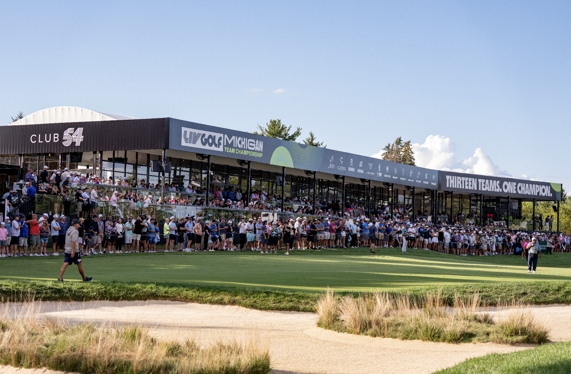 Spectators watching a golf tournament at Club 54, with a large digital banner displaying event details and various sponsor logos, against a blue sky with some clouds.