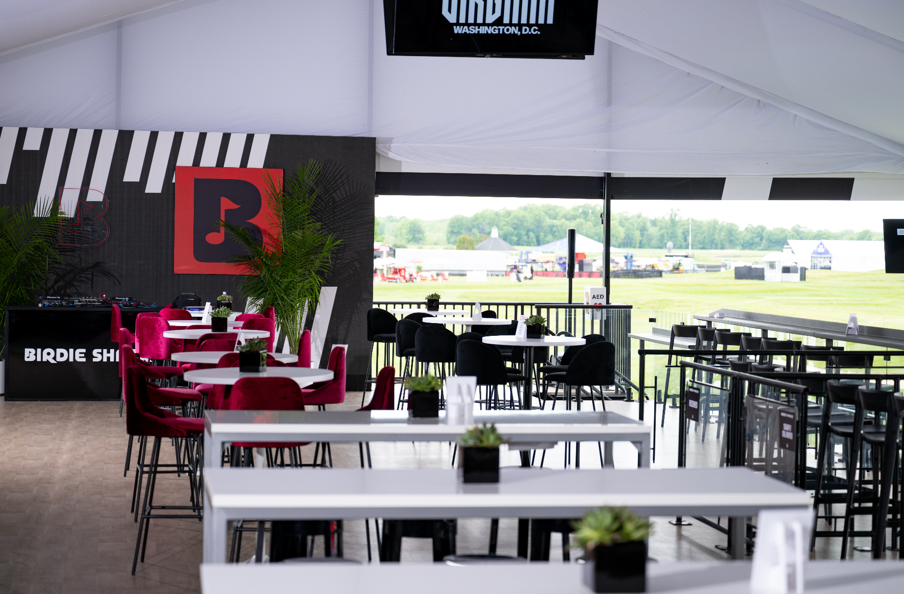 Interior of a modern event tent with chairs and tables, overlooking an outdoor field with tents and equipment in the distance.