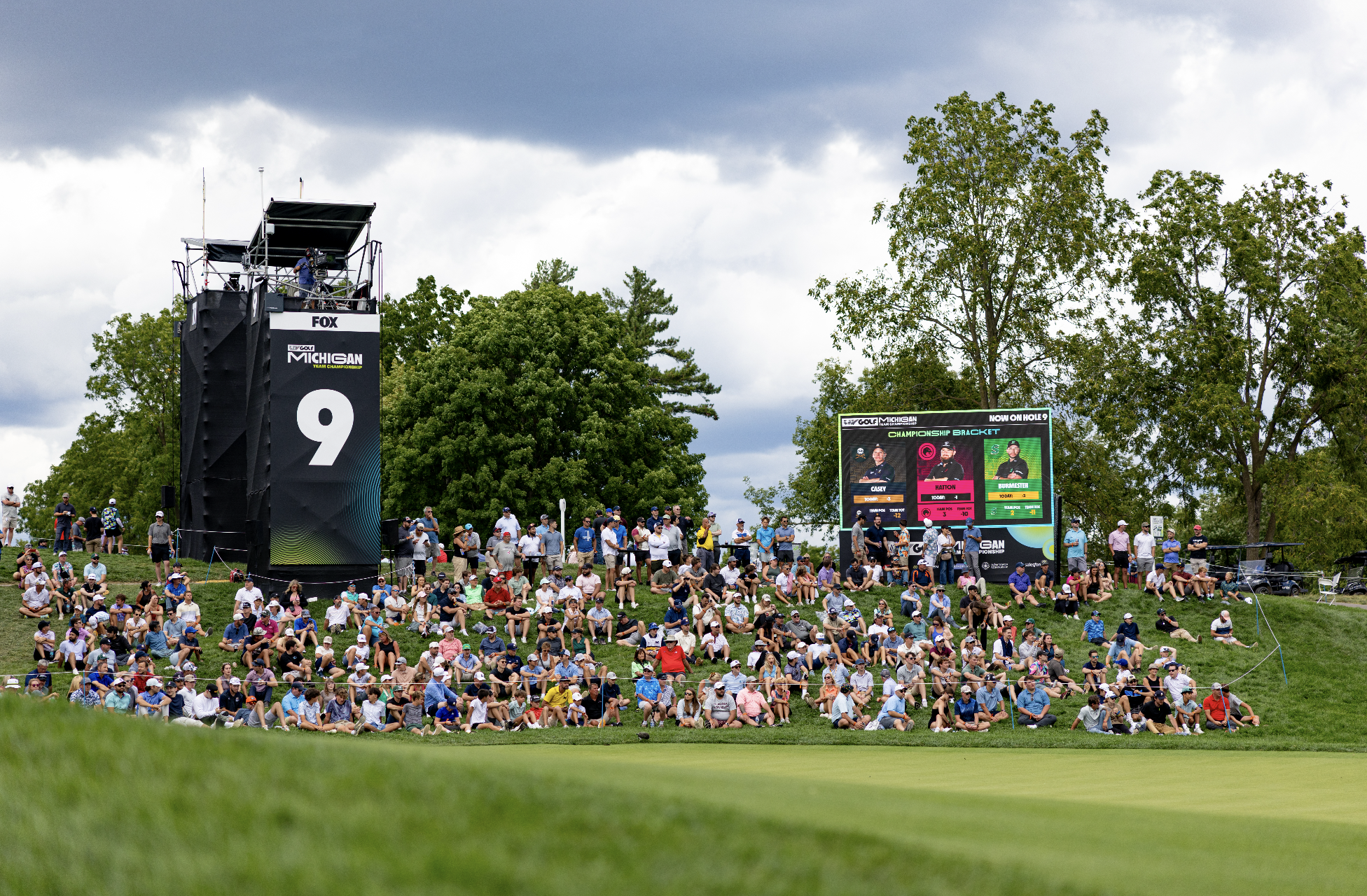 Crowd of spectators sitting and standing on a grassy hill watching a golf tournament, with a large digital scoreboard and a tower with a camera on top under a cloudy sky.