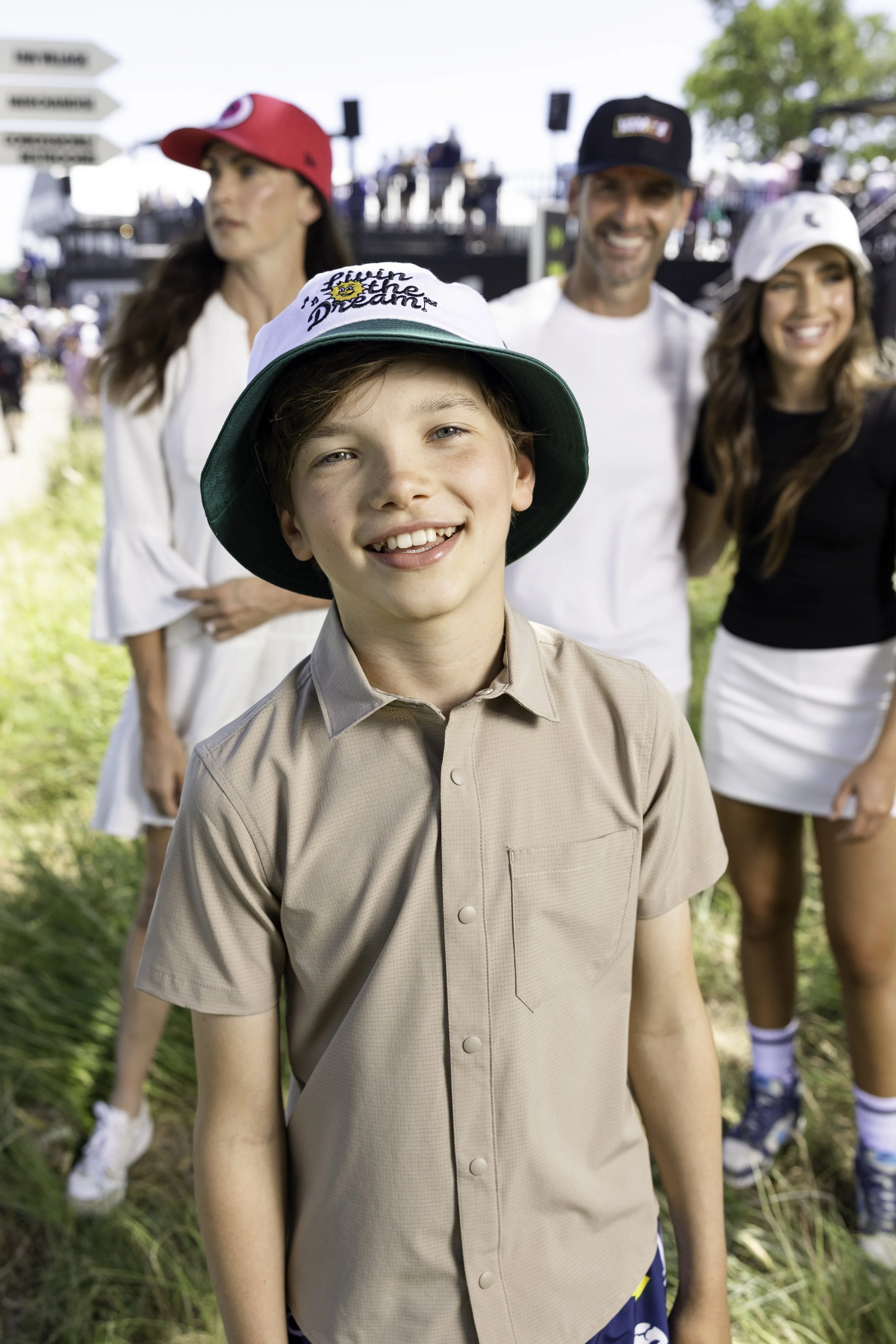 Four people standing outdoors on a sunny day, with the focus on a young boy in the foreground wearing a tan short-sleeved shirt and a white hat that says "Dreamer". Behind him are a woman with long dark hair wearing a white blouse and a red cap, a smiling man with a beard wearing a black cap and a white t-shirt, and another woman with long wavy hair wearing a white hat and a black shirt. There are trees and a social event setup in the background.
