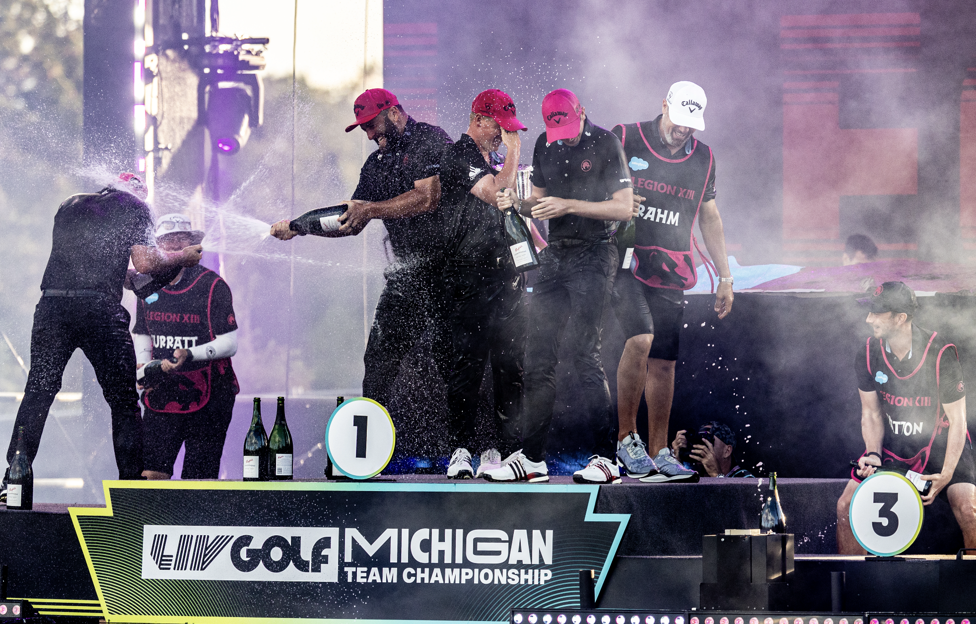 A group of four male golfers celebrating on a winners' podium at the LIV Golf Michigan Team Championship, with bottles of champagne spraying and standing near a sign indicating the first-place position.
