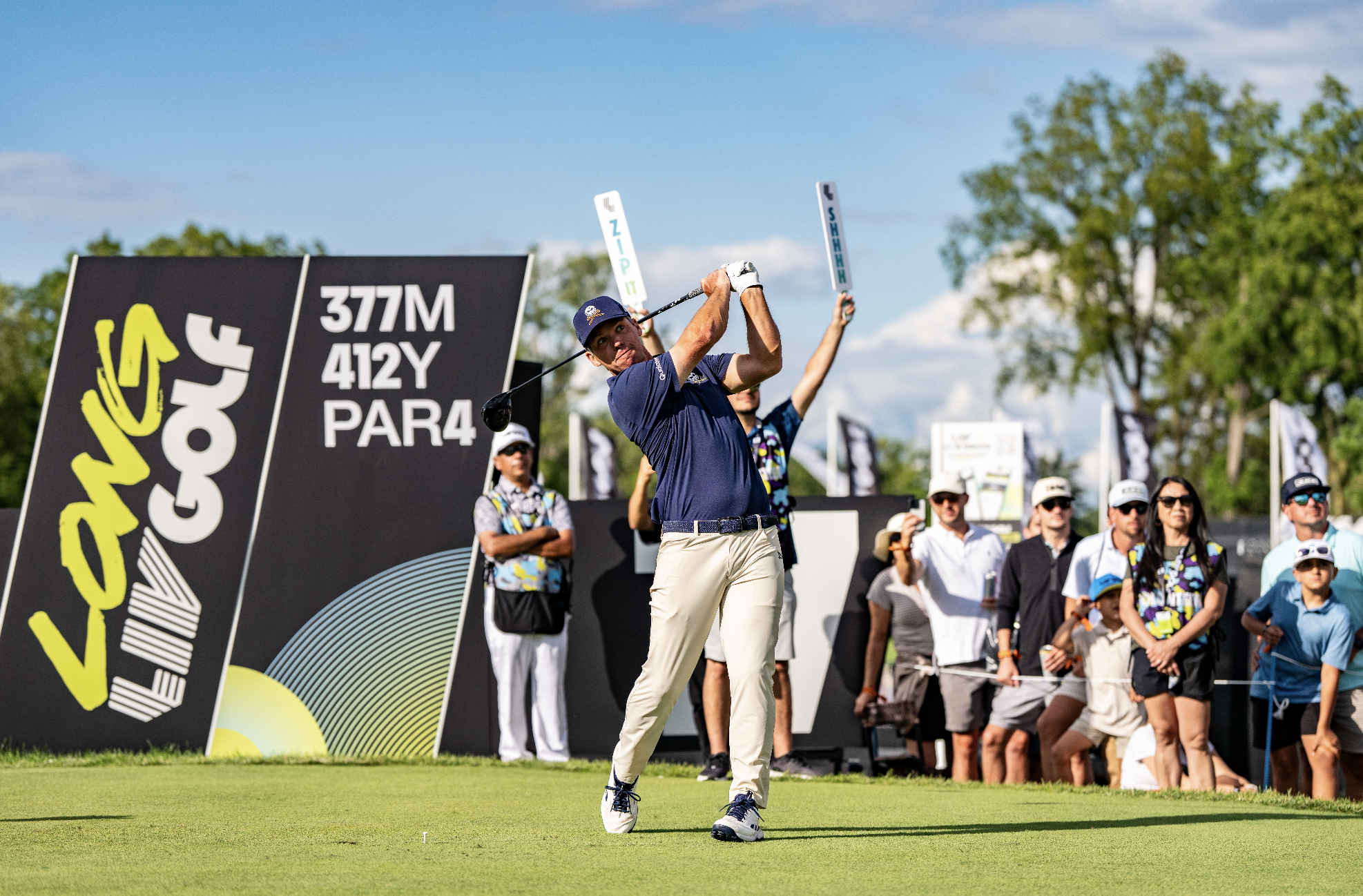 A golfer swings a club on a bright golf course with a blue sky, trees, and a crowd of spectators watching. There are signs with distances and game information in the background.