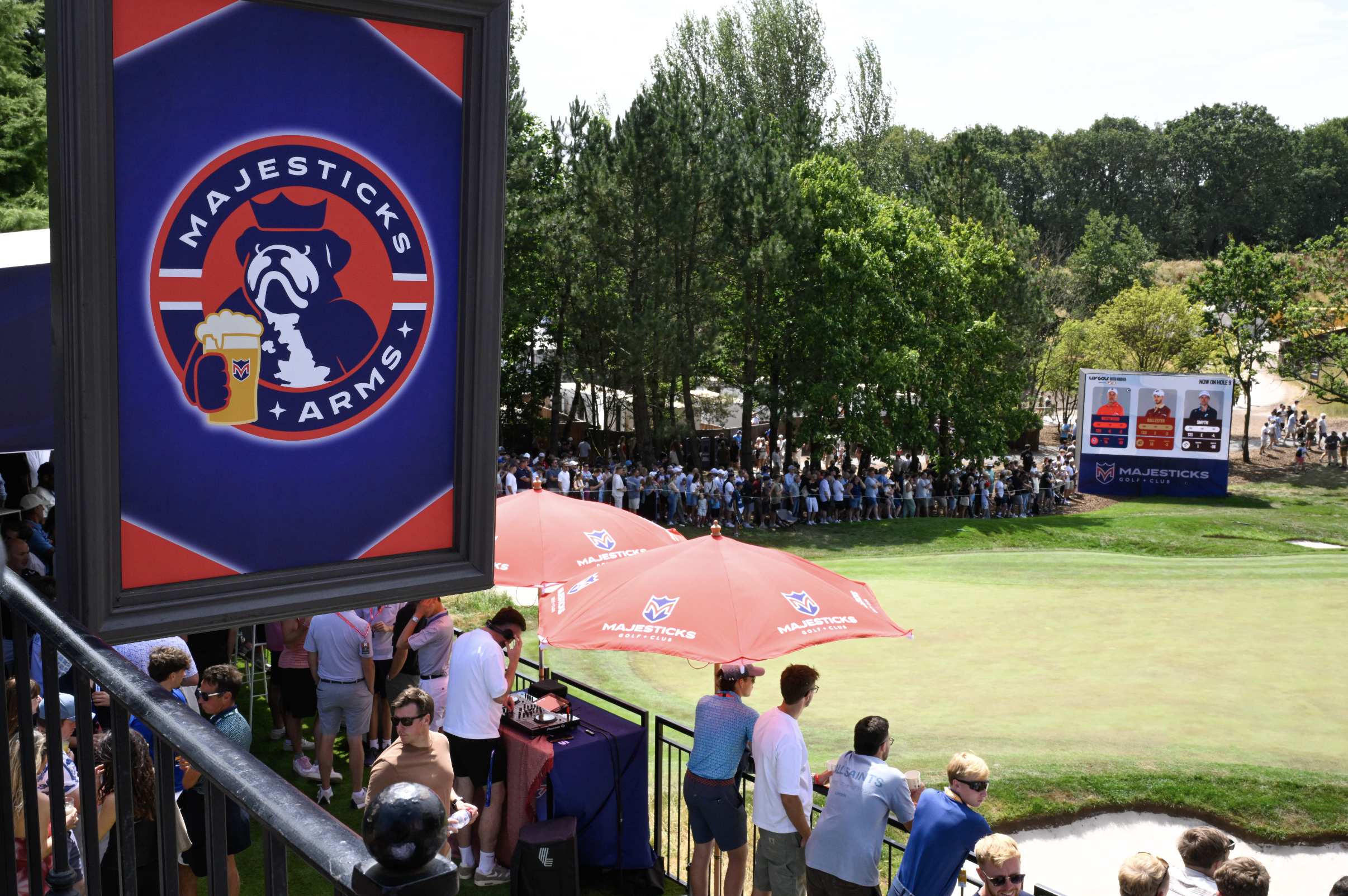 A crowd of people gathered at a golf course during a tournament, with a Majesticks Golf Club sign in the foreground and a scoreboard in the background showing scores of golf players, all surrounded by green trees.
