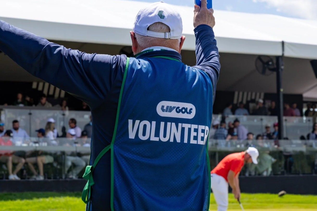 A man wearing a white cap and a blue vest with a green strap, which has the word 'VOLUNTEER' on the back, stands on a golf course with arms raised. In the background, there are people sitting in a balcony and a person in an orange shirt preparing to swing a golf club.