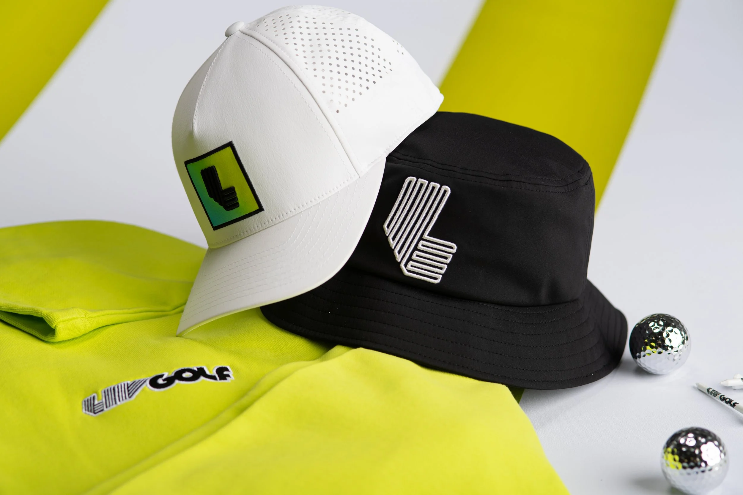 A collection of golf accessories including three caps in black, white, and black with white logos, a neon yellow golf shirt with black and white text, a neon yellow hat, and small golf balls with black and yellow patterns, all placed on a white surface with a yellow background.