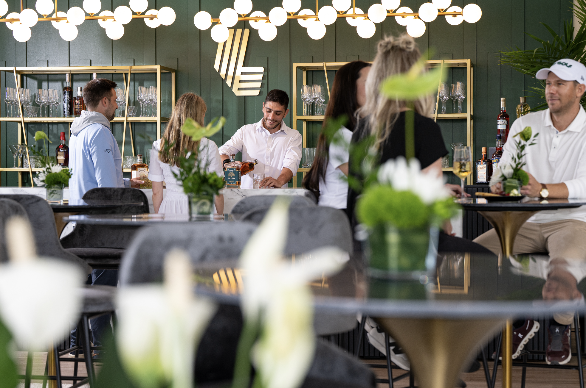 People socializing and enjoying drinks at a modern bar or restaurant with dark green walls, bright lighting, and gold accents, with flowers and plants in the foreground.