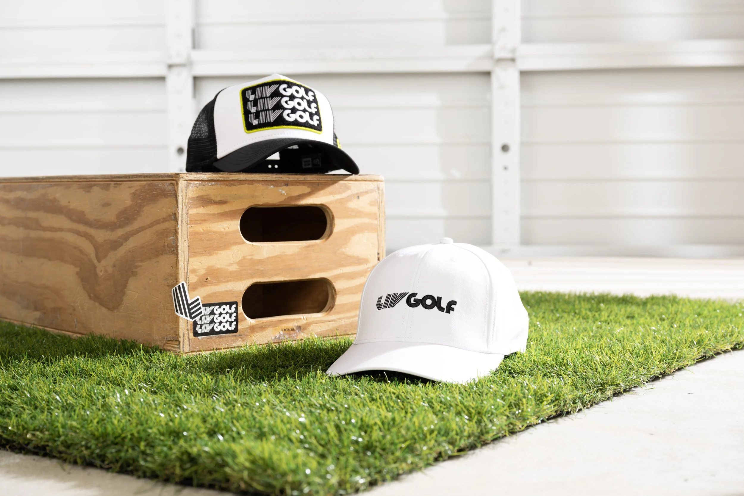 A white golf cap with the Living Golf logo on it, resting on a patch of green grass next to a wooden box with the same logo, with a white background wall.