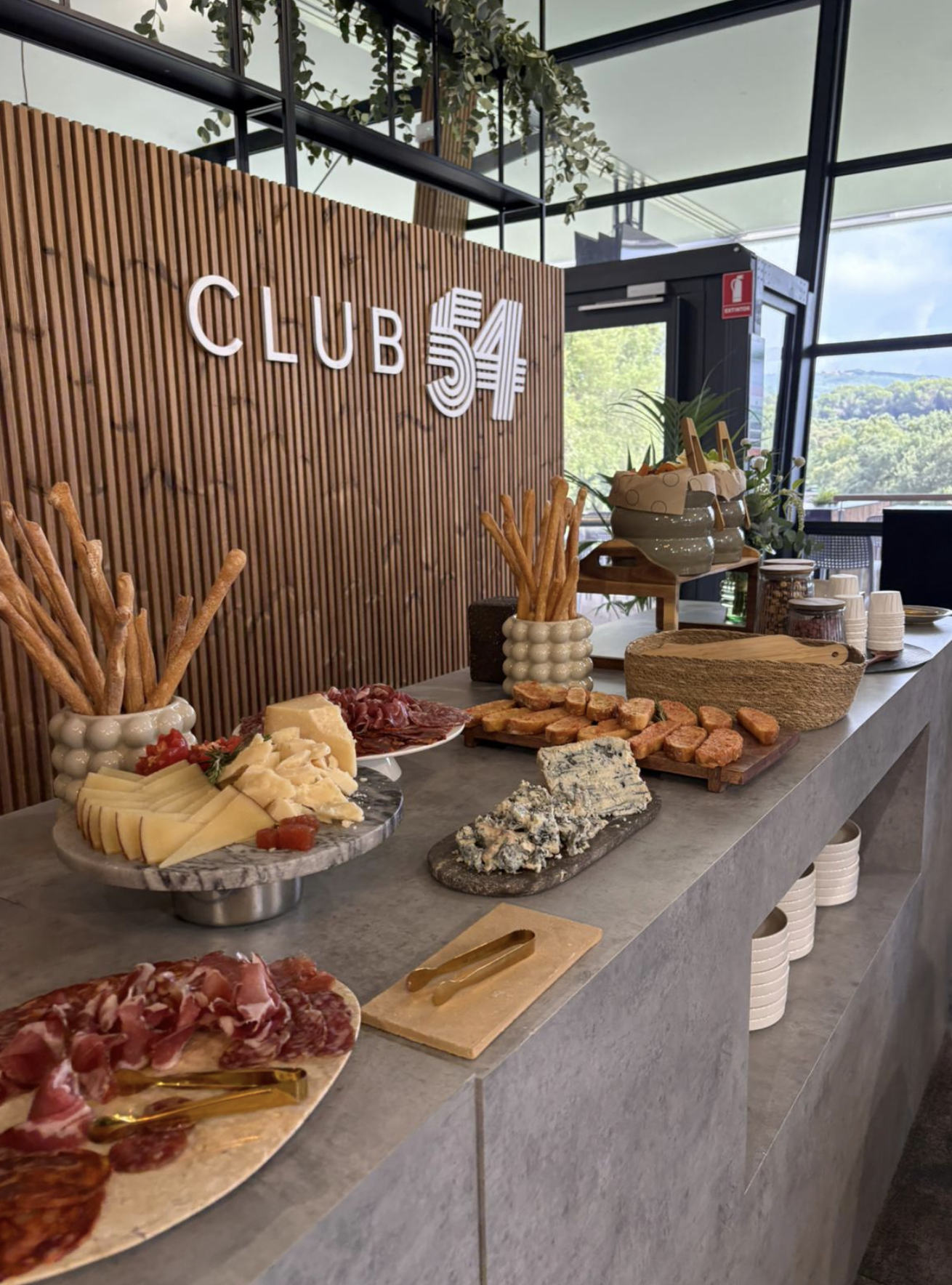 Charcuterie and cheese platter with sliced cured meats, cheeses, breadsticks, and crackers on a marble table with a wooden backdrop and the sign 'CLUB 54' in the background.