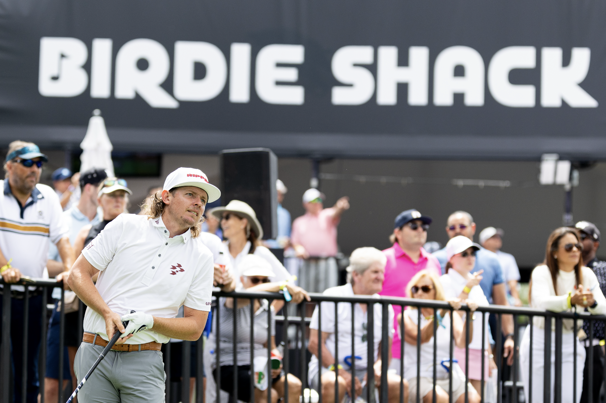 A golfer with long hair and a white shirt holding a golf club, standing near a crowd of spectators at a golf tournament, with a large sign reading 'BIRDIE SHACK' in the background.