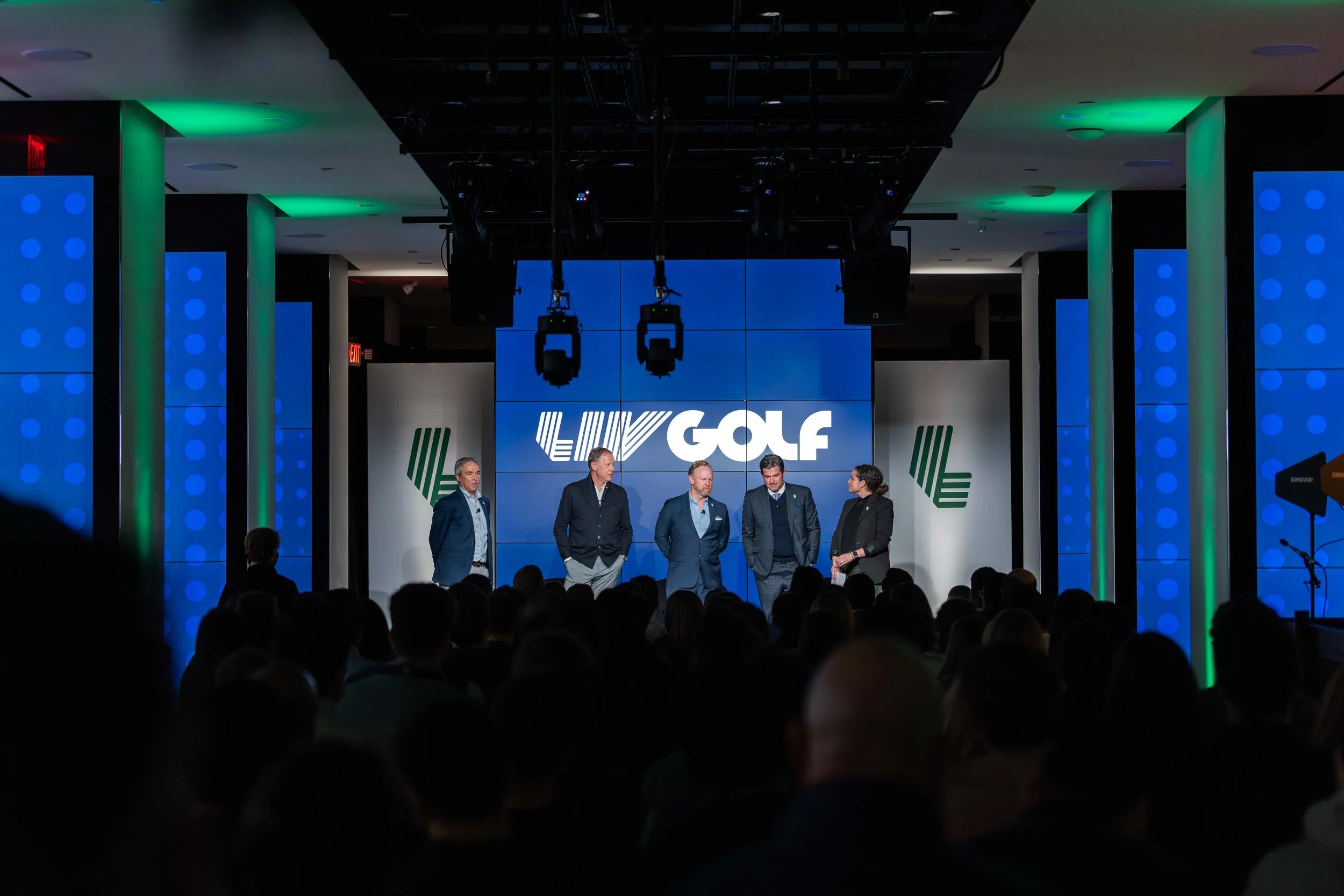 A group of five people standing on a stage in front of a large screen displaying the words 'LIV GOLF' at an indoor event with audience members visible in the foreground.