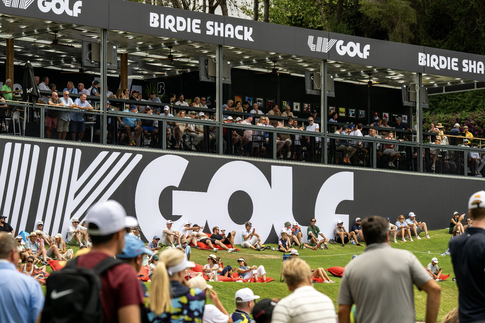 People sitting and standing at a golf tournament during the day, watching from grassy areas and a viewing deck with signs for Birdie Shack and LIV Golf.