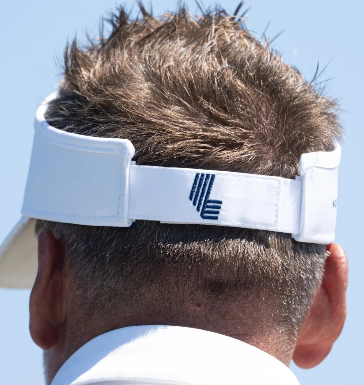 Back of a person's head with brown hair, wearing a white cap with navy blue logo, against a clear blue sky.