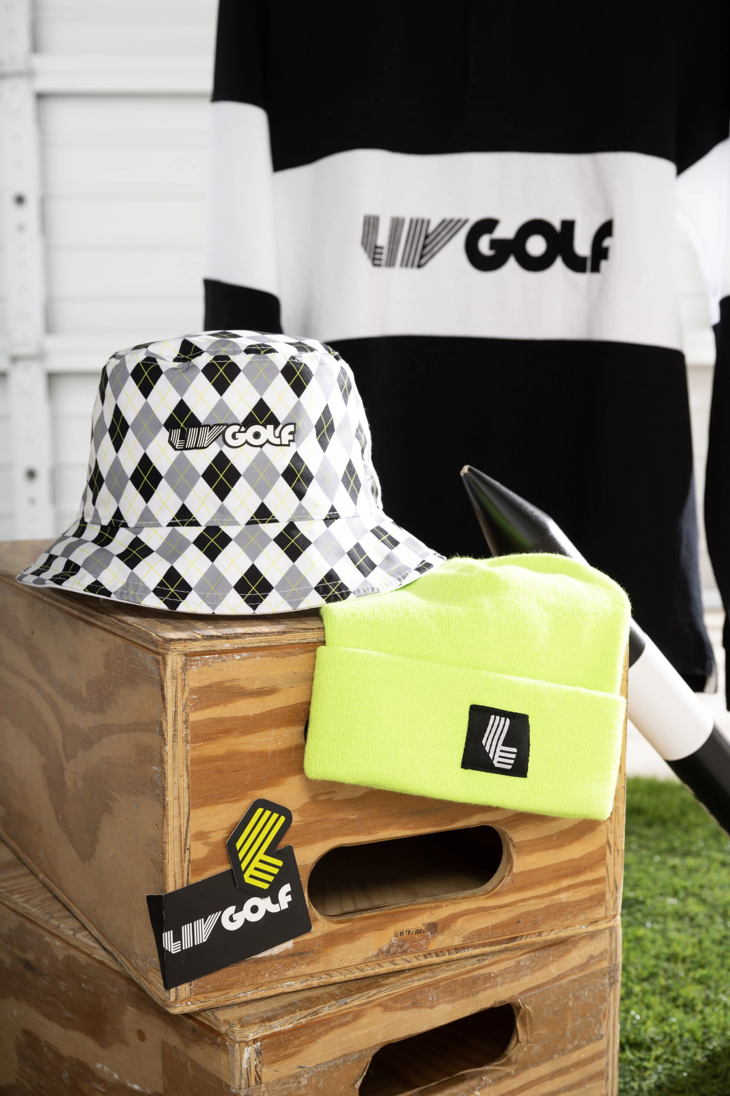 Golf-themed merchandise on a wooden display at an outdoor event, including a plaid golf hat, a neon yellow beanie, and apparel with branding.