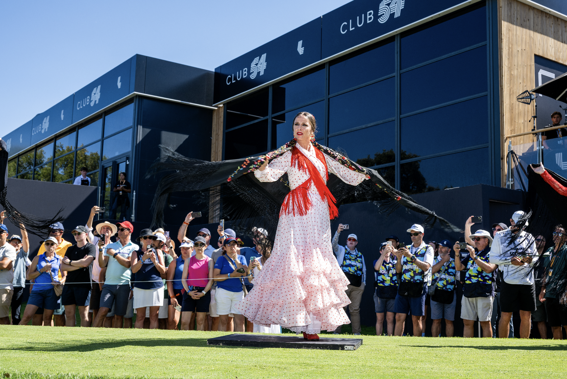 A woman in a traditional Spanish dress performing a flamenco dance on a stage at a golf tournament, with a crowd of spectators watching.