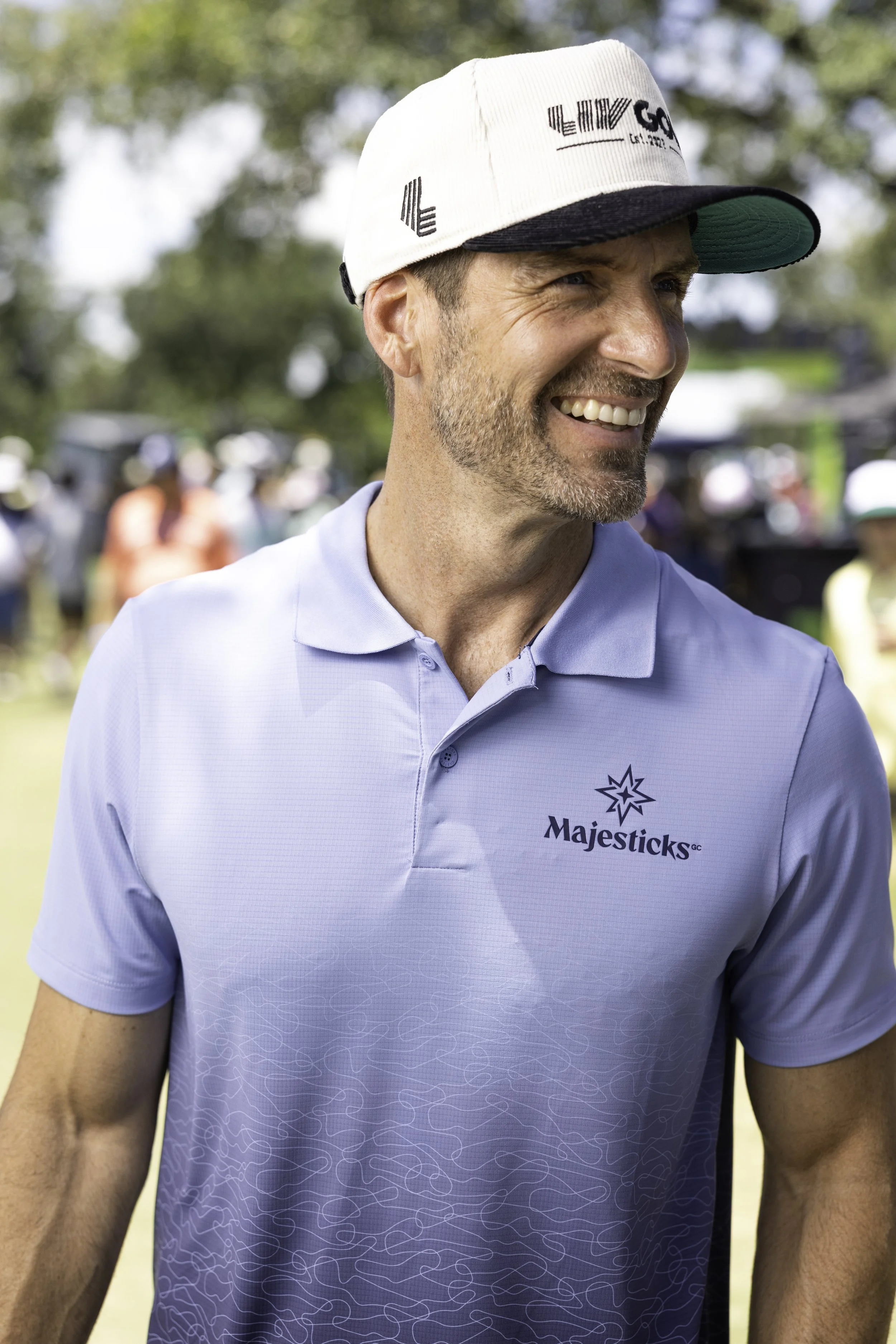 Close-up of a man wearing a light blue polo shirt with a logo that reads "Majesticks GC" and a white and black baseball cap, smiling outdoors with a blurred background of trees and people.