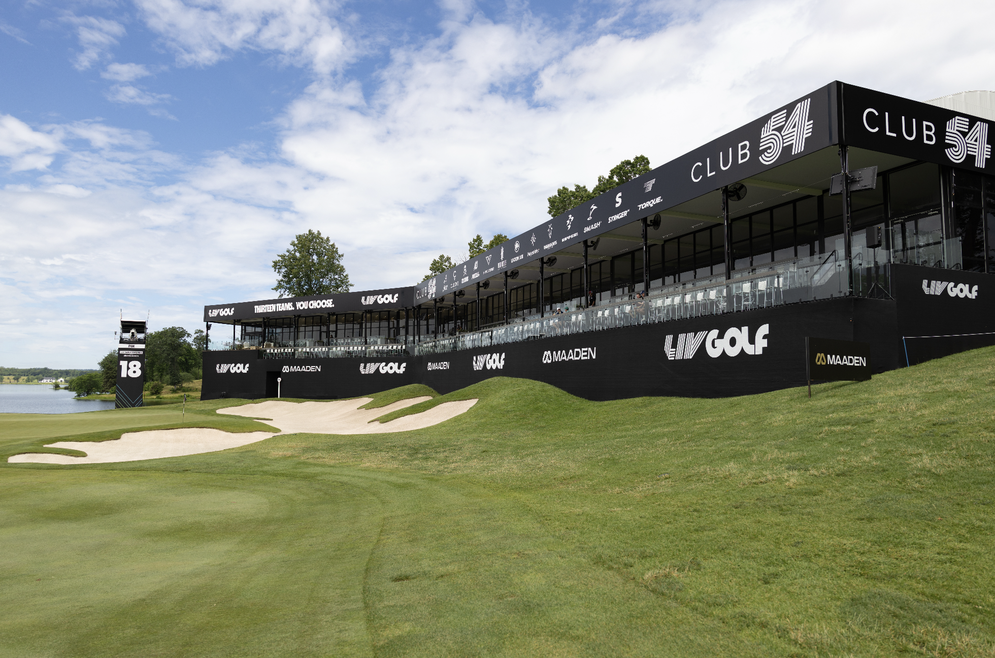 A golf course with a large viewing stand labeled 'Club 54' and various sponsors, overlooking green fairways and sand traps, under a partly cloudy sky.