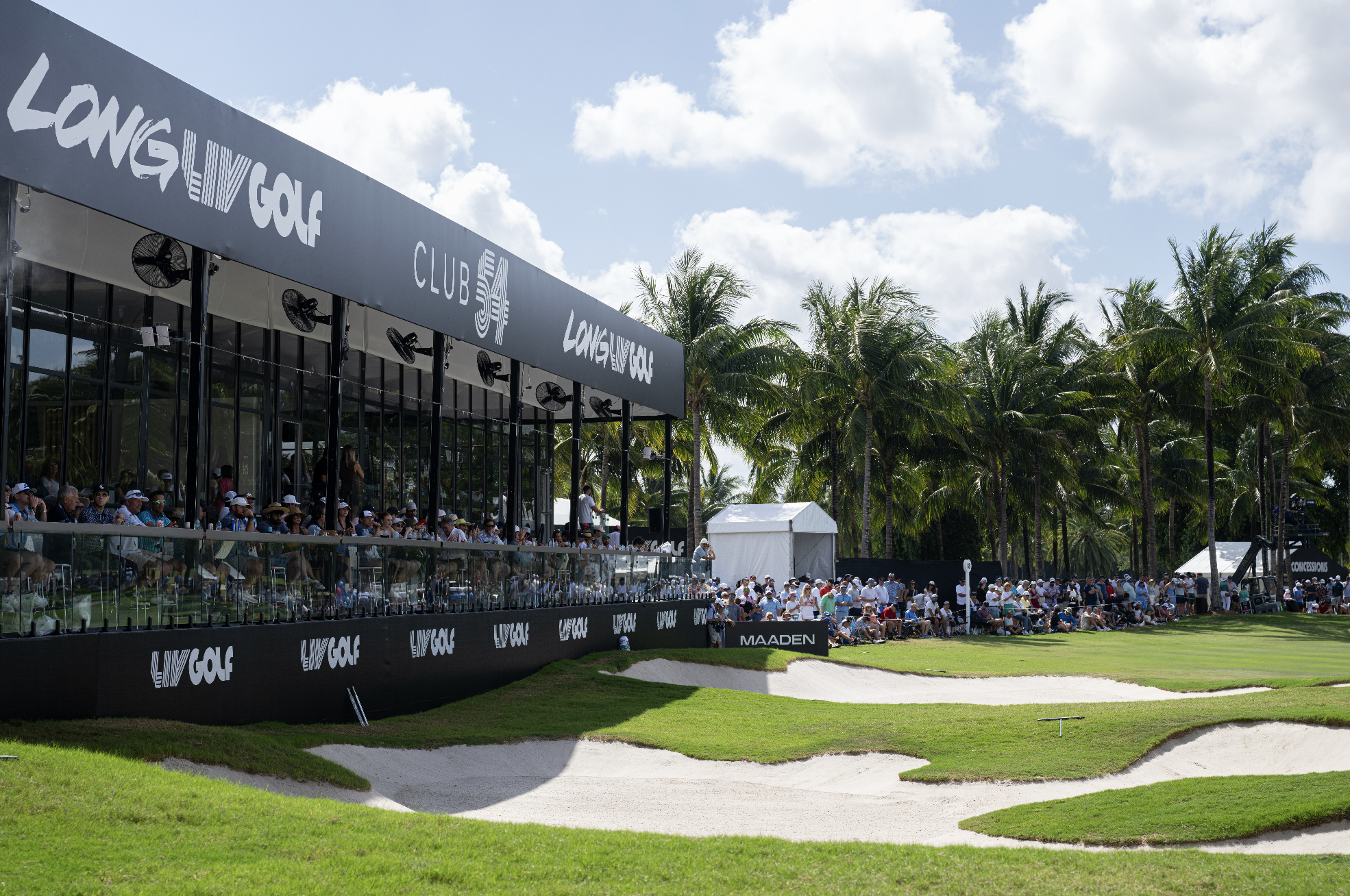 Crowd watching a golf tournament at a tropical golf course with palm trees and blue sky