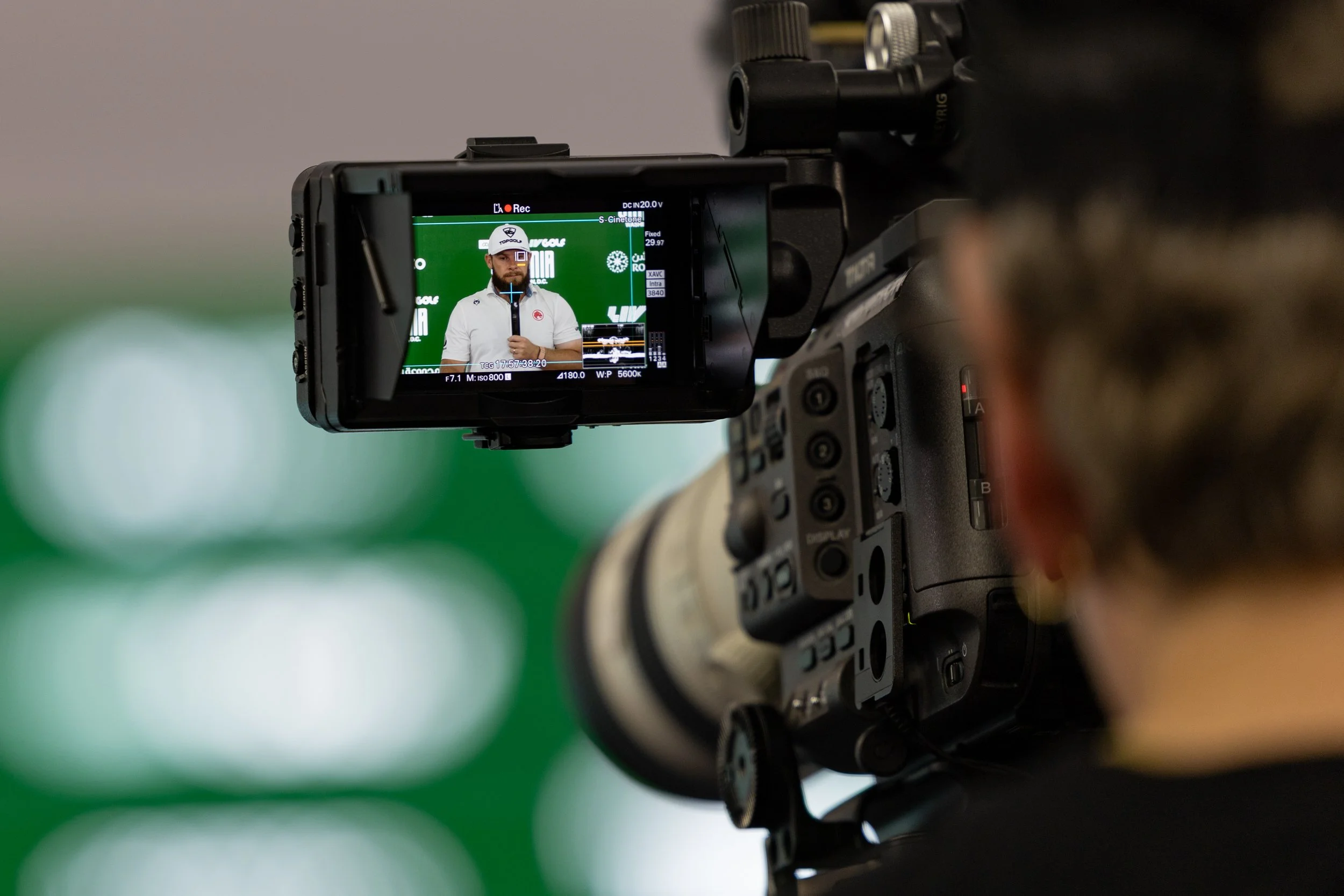 A person is filming a man holding a microphone in front of a green background with sponsor logos.