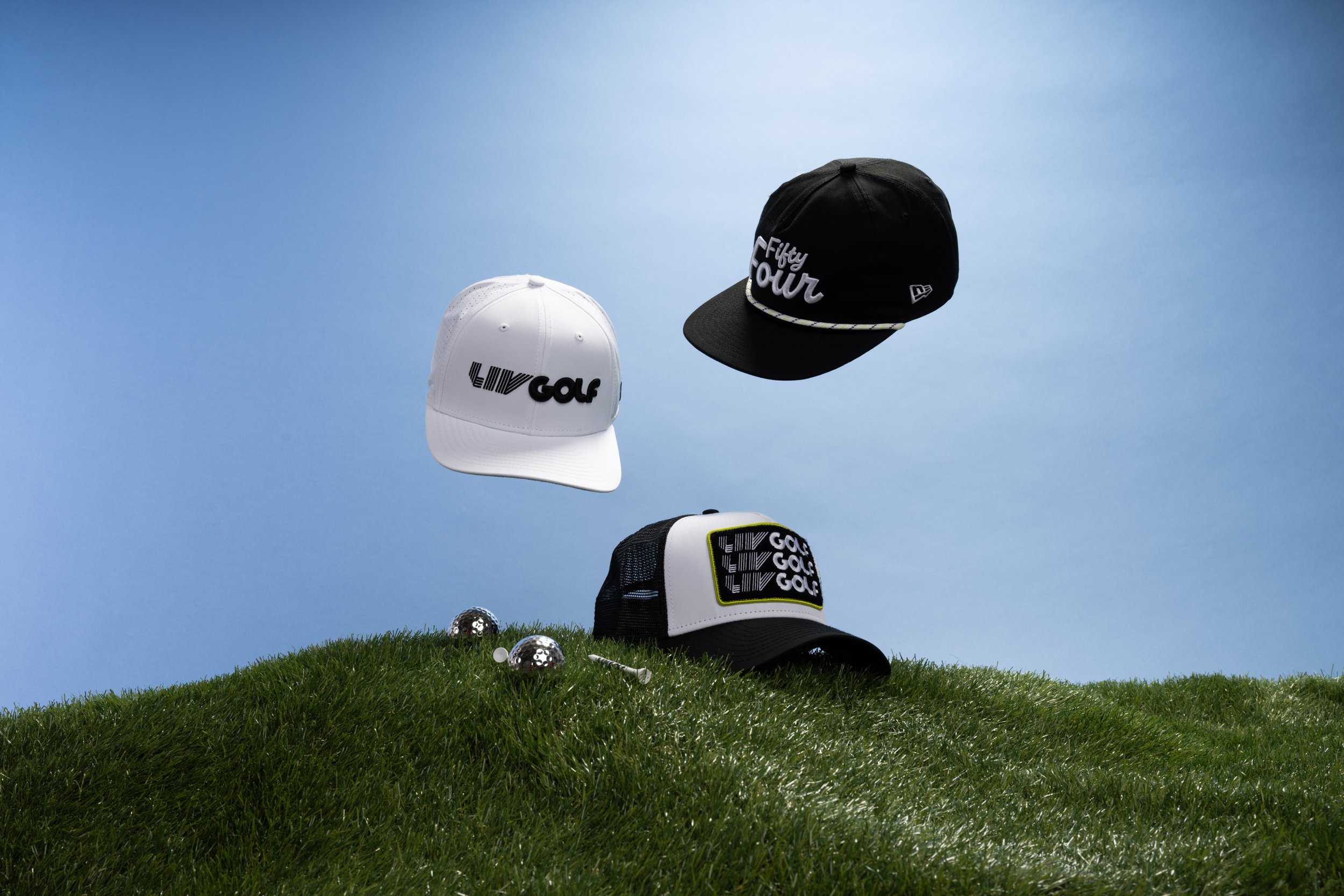 Four golf caps placed on a grassy hill with a clear blue sky background. One white cap with the text 'LIVE GOLF' and two black caps with the texts 'Fifty Four' and a pattern. Several metallic golf balls are on the grass.