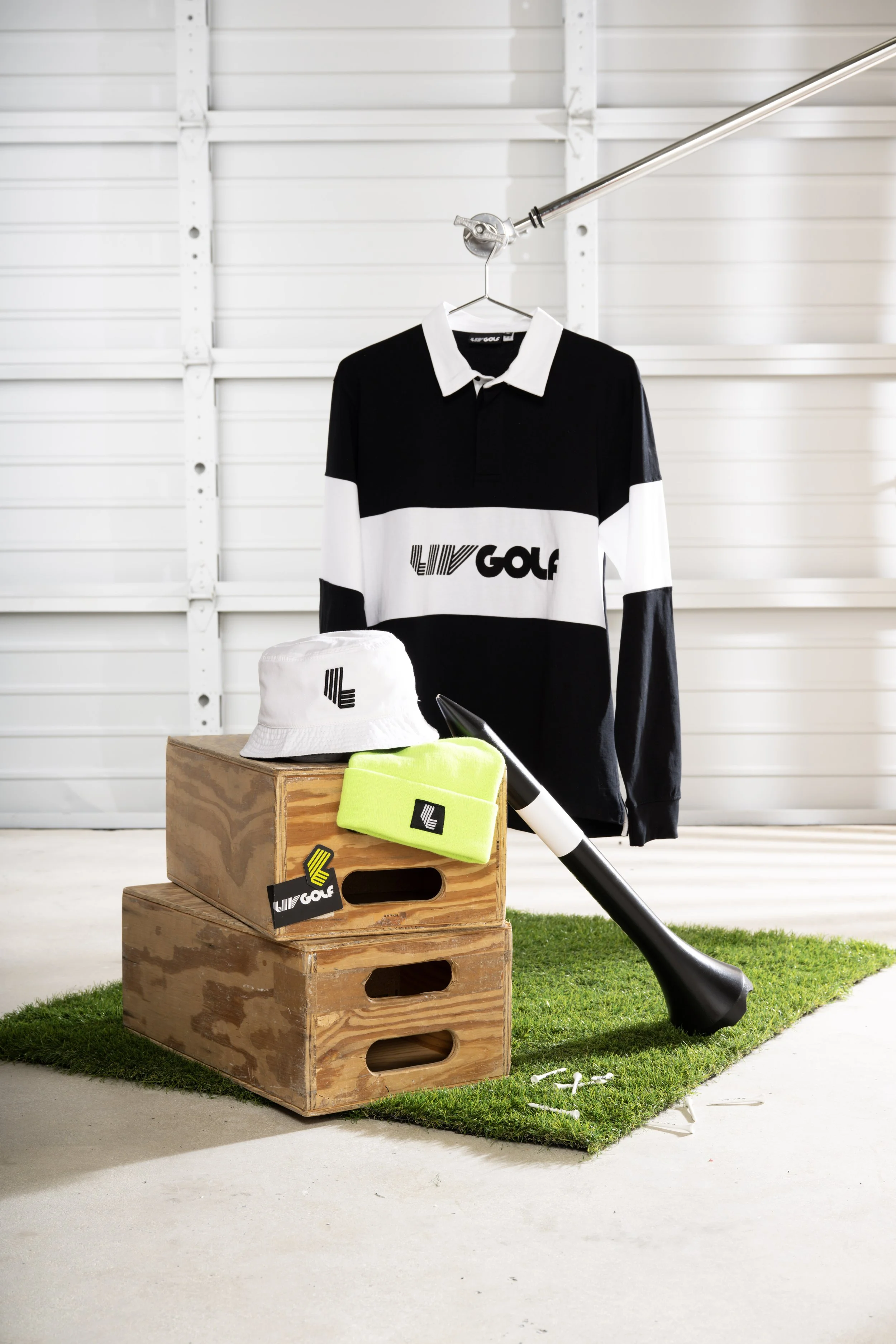 A golf-themed display with a black and white LIV GOLF shirt hanging on a hanger, a white LIV GOLF bucket hat, yellow LIV GOLF towel, and white LIV GOLF tags, all arranged on a wooden crate with a golf club lying on the ground nearby, in a setting with a white garage door in the background and synthetic turf underfoot.
