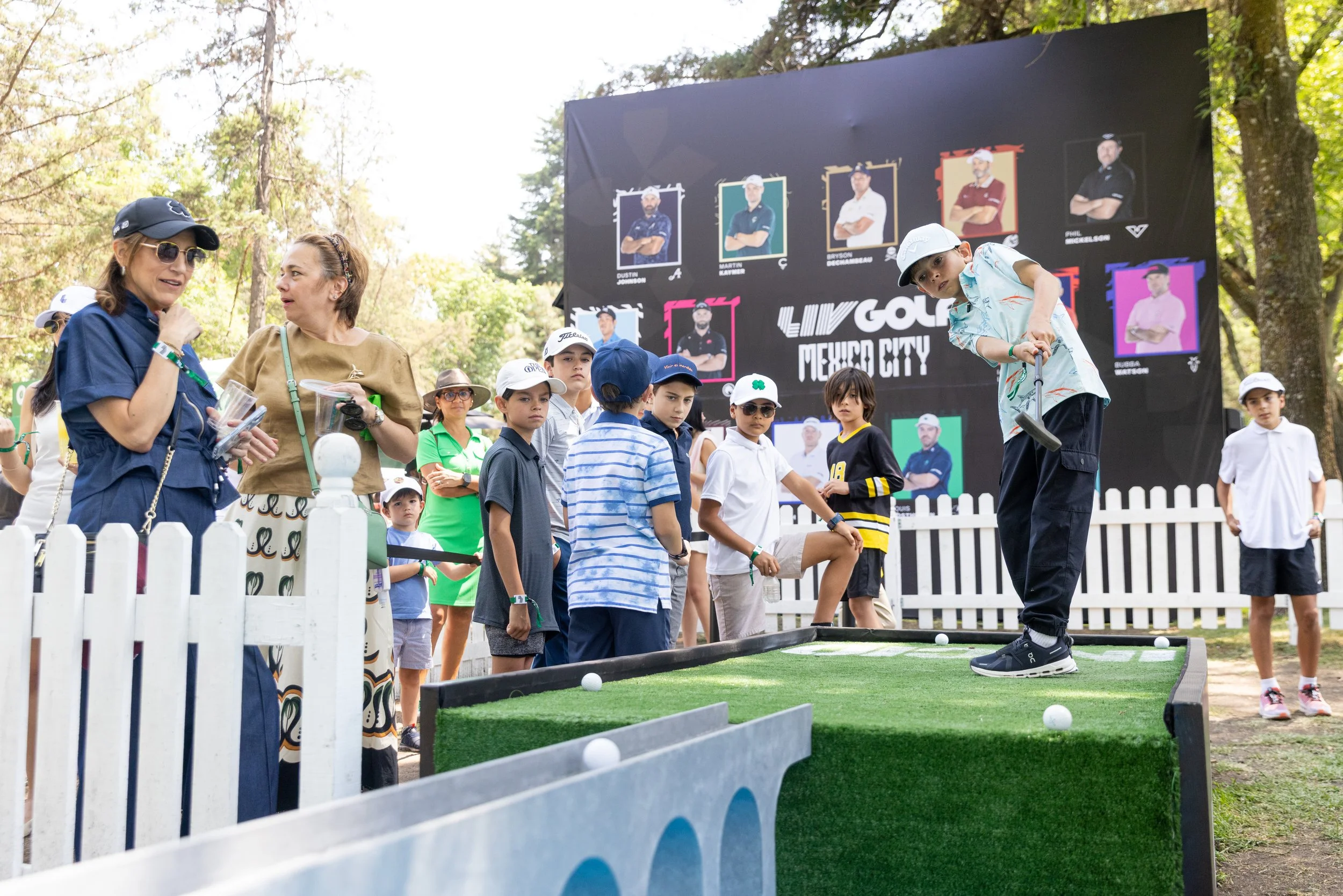 Kids and adults gathered outdoors at a mini-golf course with a large digital leaderboard in the background, displaying player photos and names.