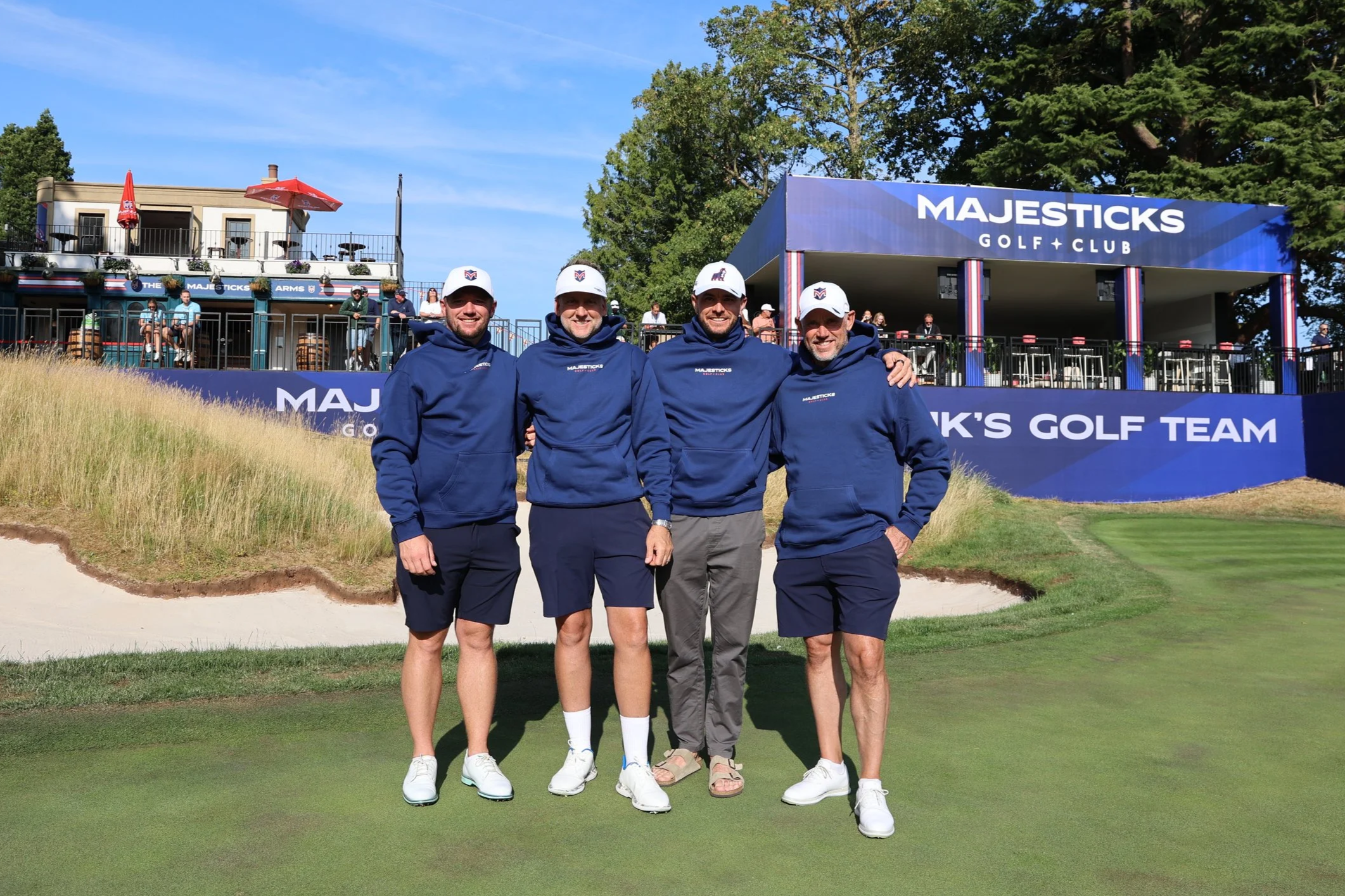 Four men standing on a golf course green, smiling at the camera, at the Majesticks Golf Club, with a clubhouse and spectators in the background.