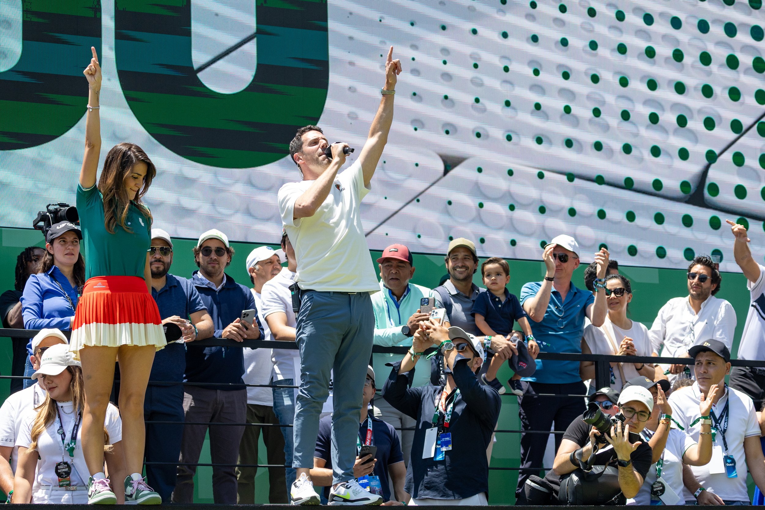 A crowd of people standing and cheering at an outdoor event, with a large digital screen in the background showing a green and black design. Two individuals are prominently visible in the foreground; a man with dark hair and a beard, holding a microphone and raising his index finger, and a woman with long hair, wearing a teal shirt and a red skirt, also raising her index finger.