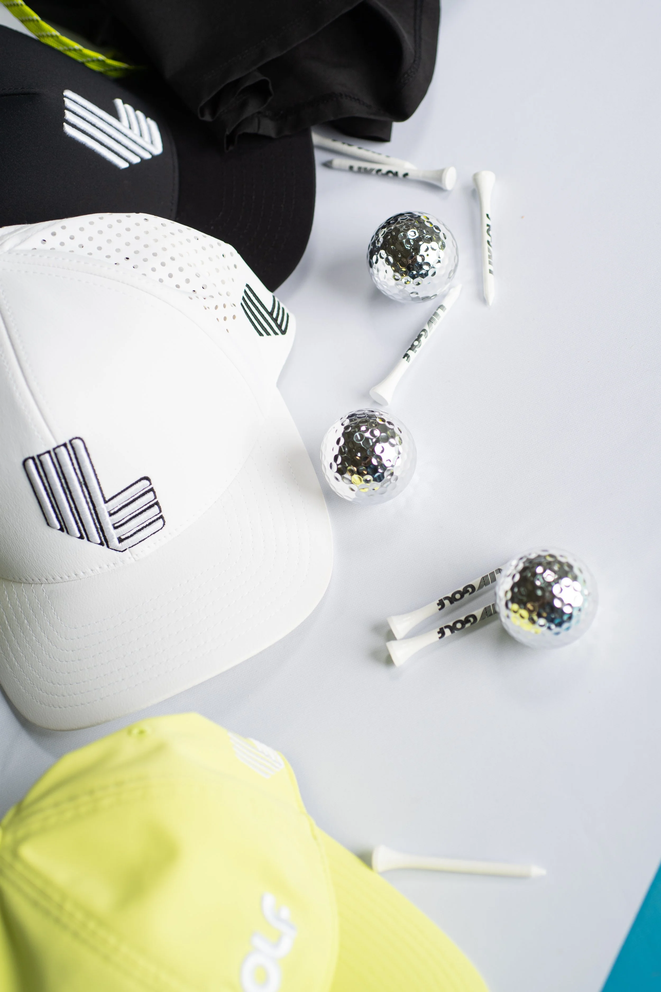 Close-up of a white baseball cap with a black logo, a black cap with a similar logo, several white golf tees with black text, reflective metallic golf balls, and part of a yellow object on a white surface.