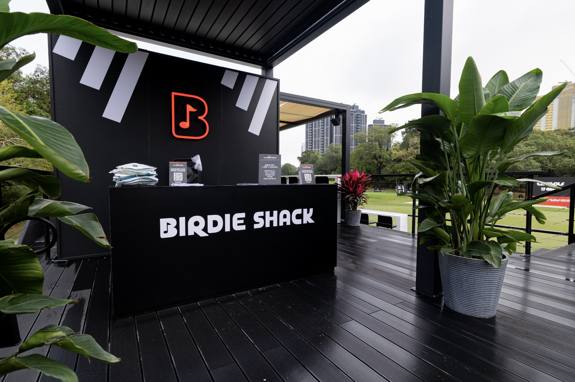 Outdoor golf event registration booth with black signage reading "Birdie Shack," surrounded by large potted green plants, with tall buildings and trees in the background.
