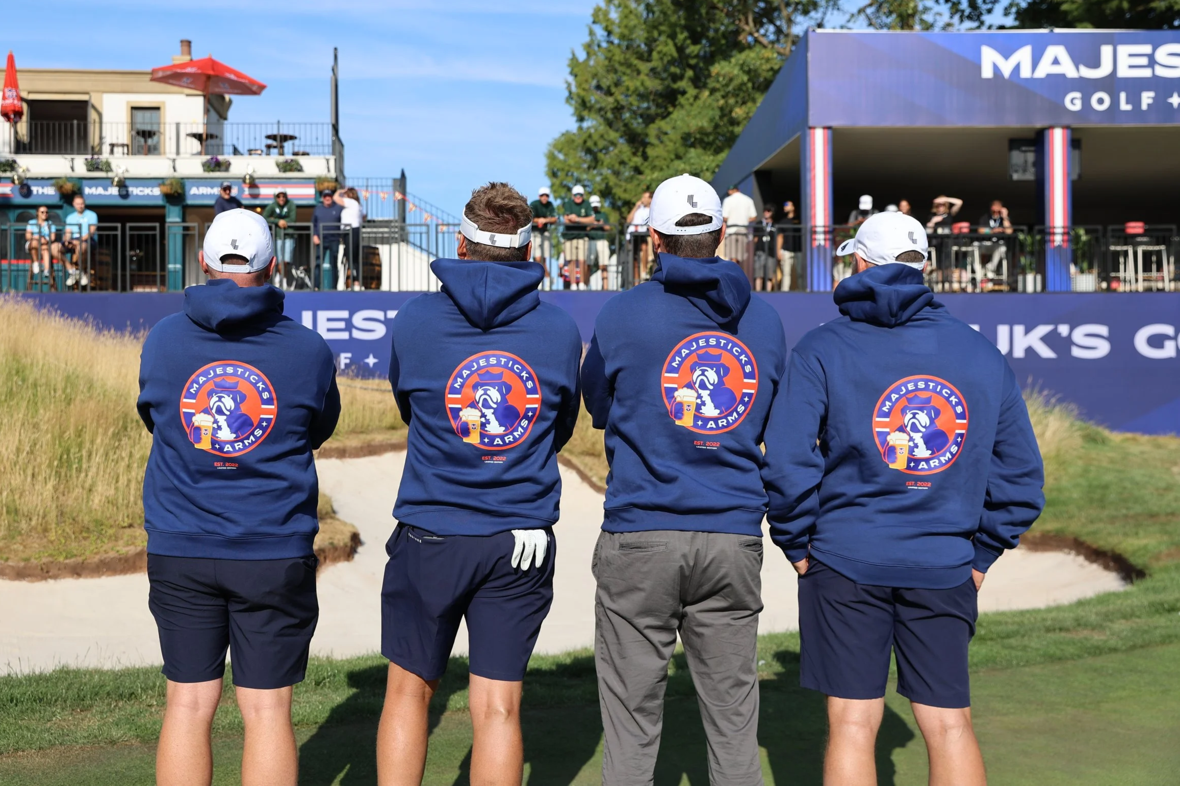 Four men walking on a golf course wearing matching navy blue hoodies with a logo on the back that says "Majesticks Arms" and featuring a bulldog holding a beer mug, with a golf club in hand.