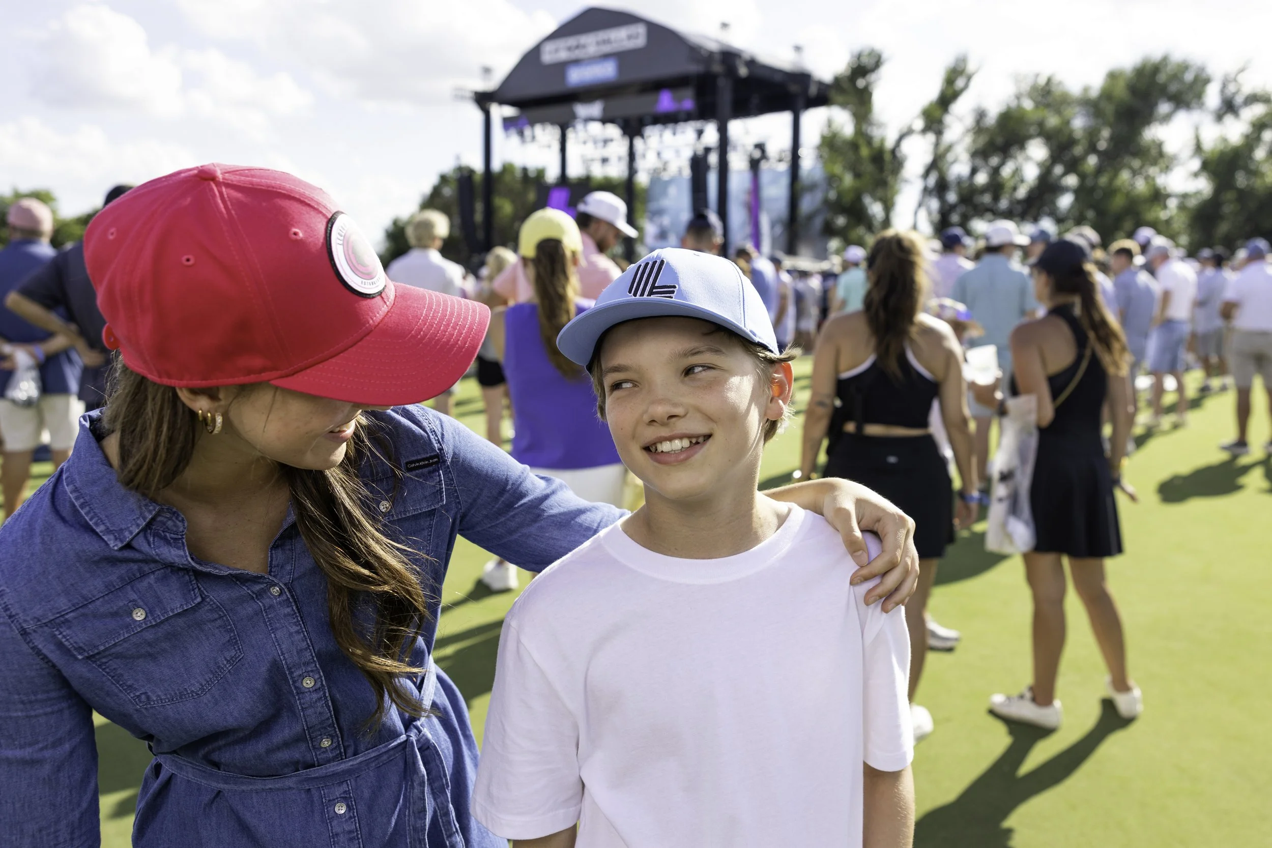 A woman and a young boy are smiling and standing close together outdoors at an event, with many people and a stage in the background.