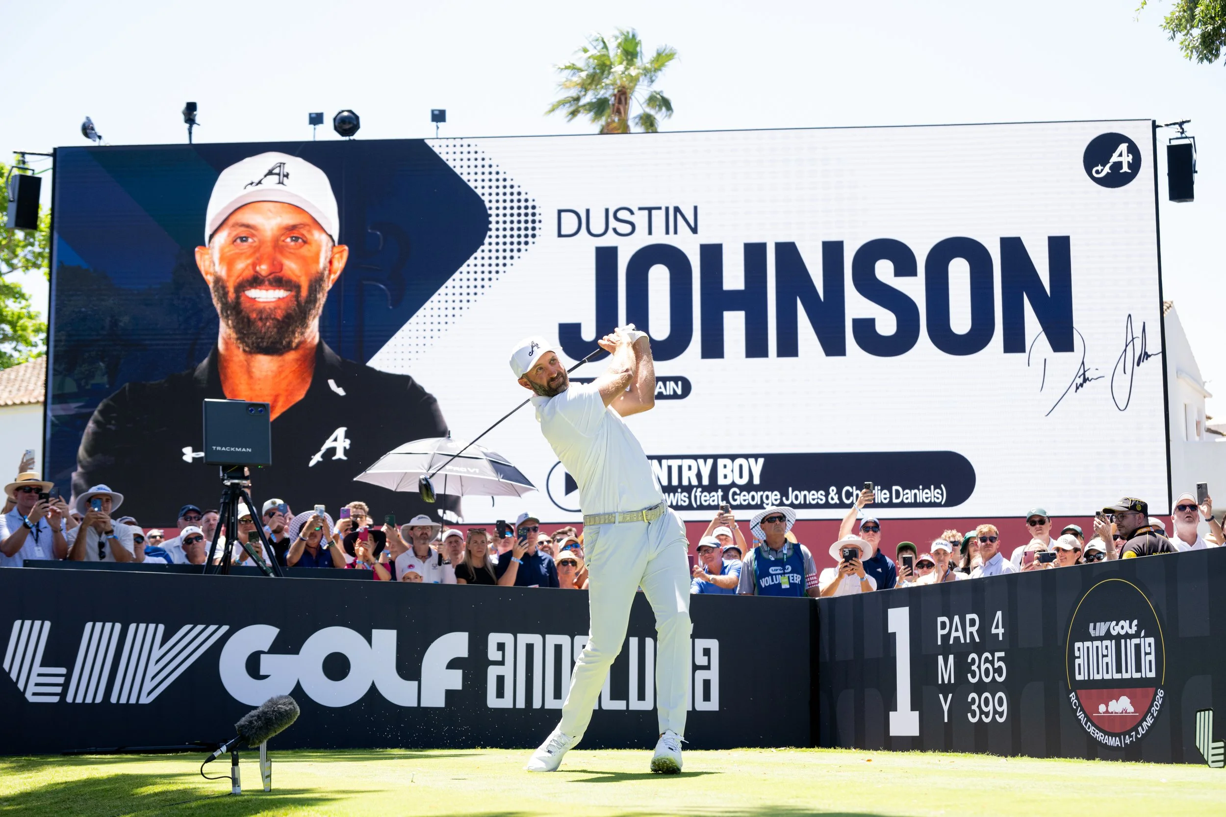 Gautam golfer swings club during a competition at a golf course, with a large digital screen displaying his name and image in the background, and a crowd of spectators watching.