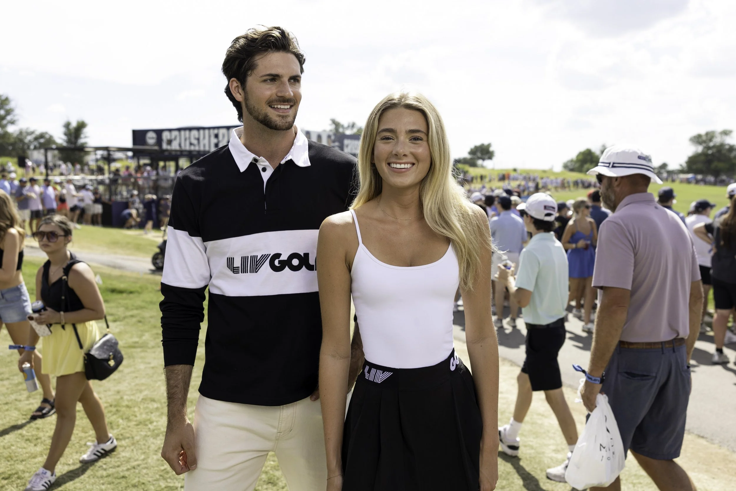 Two people, a man and a woman, smiling at an outdoor event with a crowd in the background. The man has dark hair and is wearing a black and white shirt, while the woman has long blonde hair and is wearing a white top.