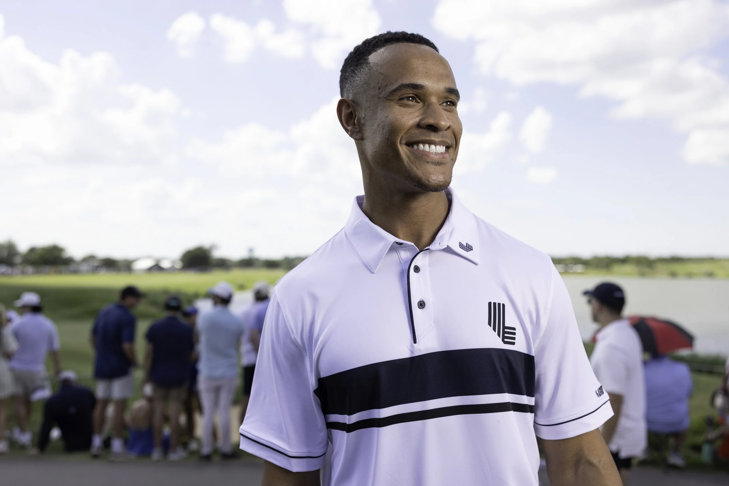 A smiling man in a white golf shirt with black and white details stands outdoors with a group of people in the background. The sky is partly cloudy.