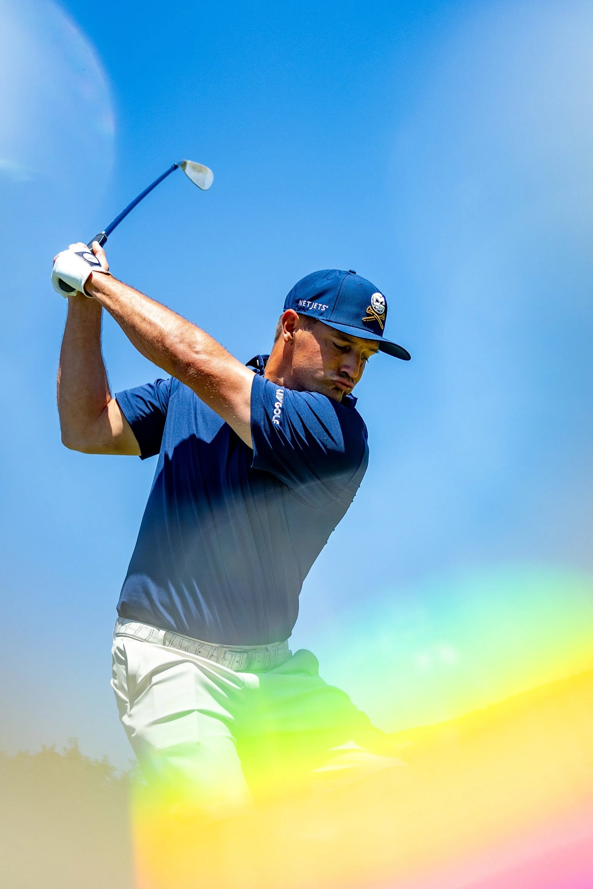 A male golfer in blue shirt and cap swinging his golf club against a bright blue sky, colorful rainbow and rainbow-colored light effects overlay the image.