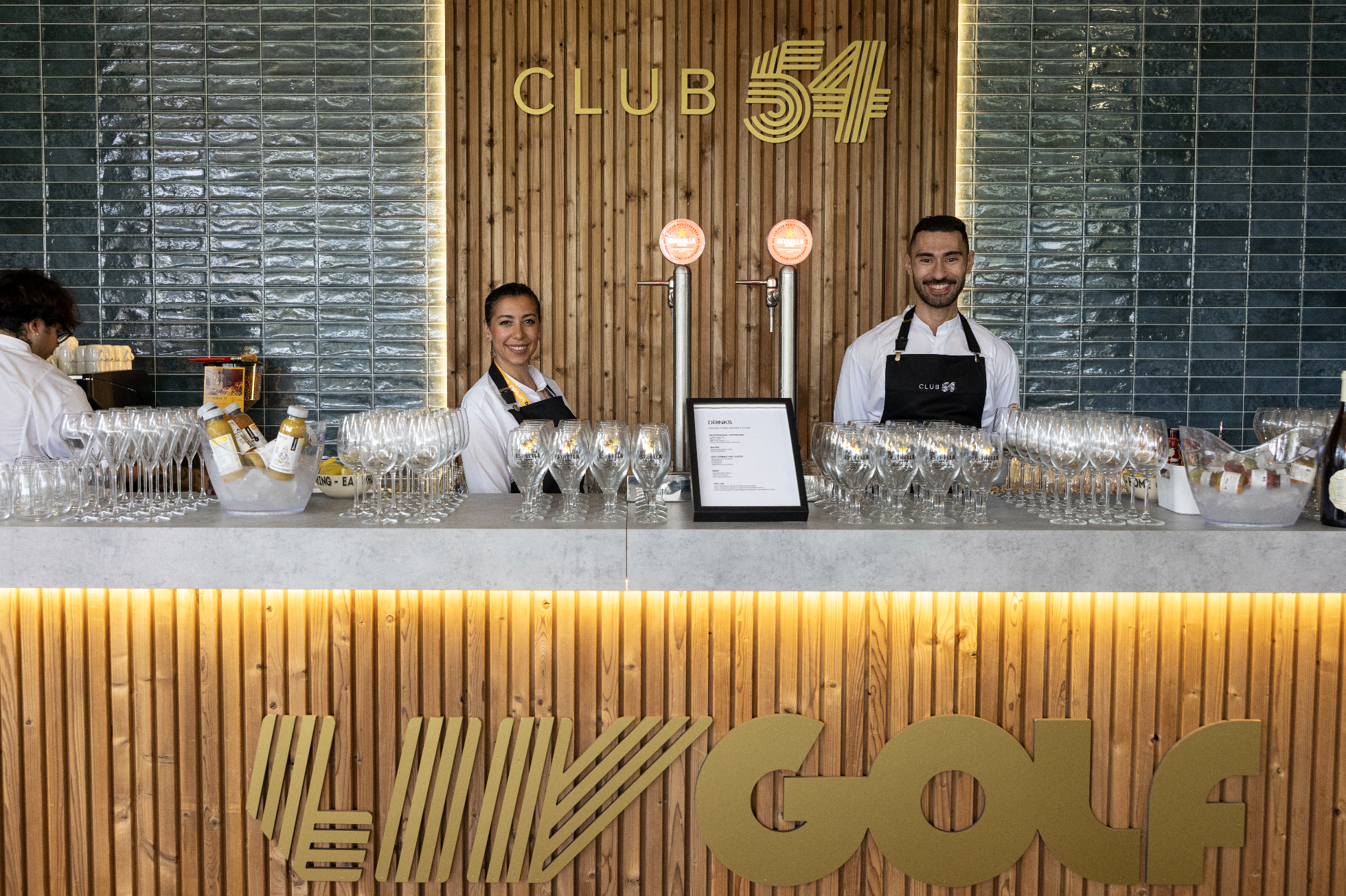 A bar at CLUB 54 featuring two smiling staff members behind the counter, with glasses and bottles on display, a menu on a stand, and a wood-paneled wall with a CLUB 54 sign illuminated by backlighting.