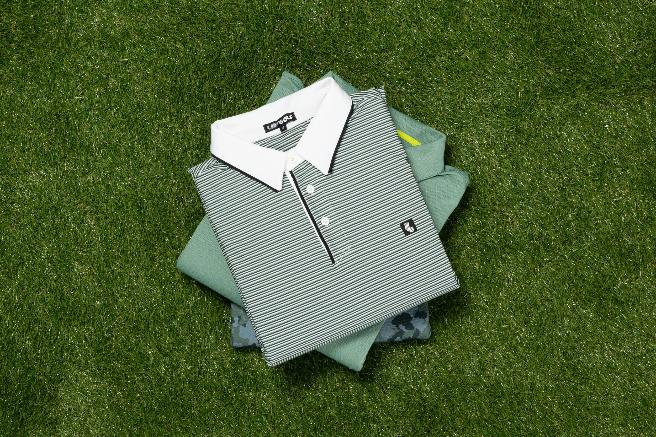 Folded golf shirts on grass, one with black and white stripes and collar, another in solid green, layered on top of a camouflaged folded item.