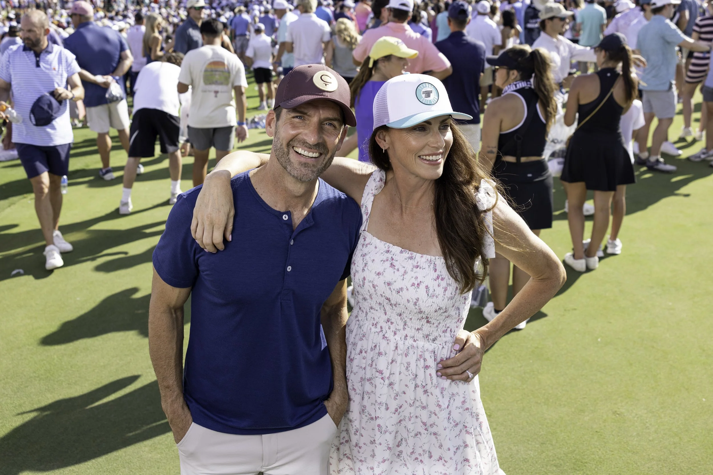 Smiling man in a maroon baseball cap and a woman in a white hat with teal accents stand together on a golf course, with a crowd of people in the background.