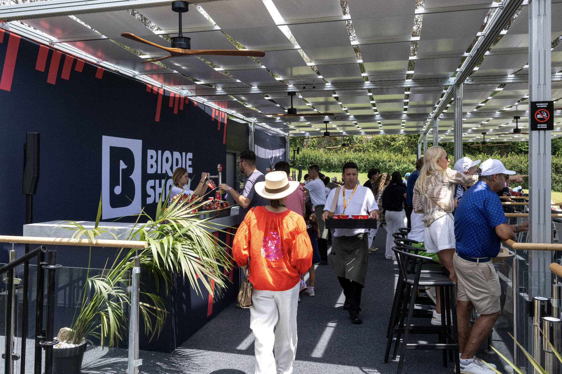 People gathered at an outdoor event at a booth with a large sign that reads 'BIRDIE SHOP.' The booth is shaded by a structure with a metal frame and solar panels on top. Several individuals are standing and interacting, some wearing white hats, and a woman in a beige patterned jacket is pointing towards something. The area has plants and outdoor seating, and the atmosphere appears lively and sunny.