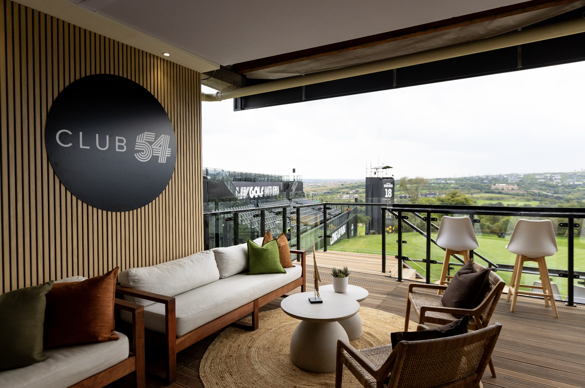 A balcony with a large round black sign reading 'Club 54' on a wood-paneled wall. The balcony features a white cushioned sofa with colorful pillows, a small white round table with a potted plant, and white and rattan chairs. Outside, there is a view of a green sports field, stadium seating, and a landscape with trees and buildings in the distance.