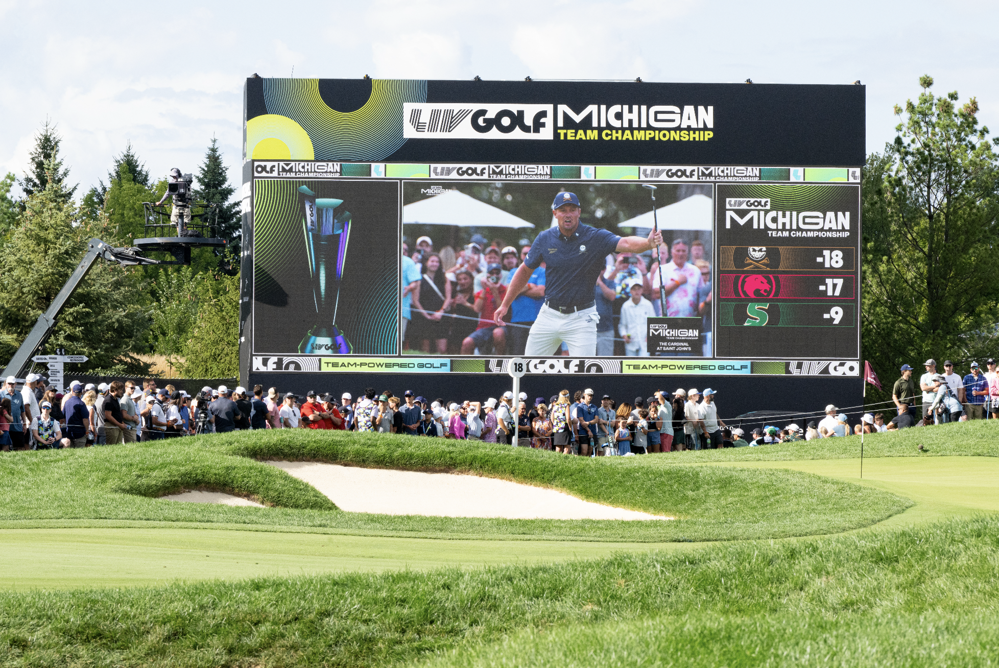 Large outdoor scoreboard displays the results of the LIV Golf Michigan Team Championship, showing team scores for three teams. The scoreboard features two images: one of a trophy and another of a golfer celebrating on the course. A crowd of spectators lines the golf course in the foreground, with trees and partly cloudy skies in the background.