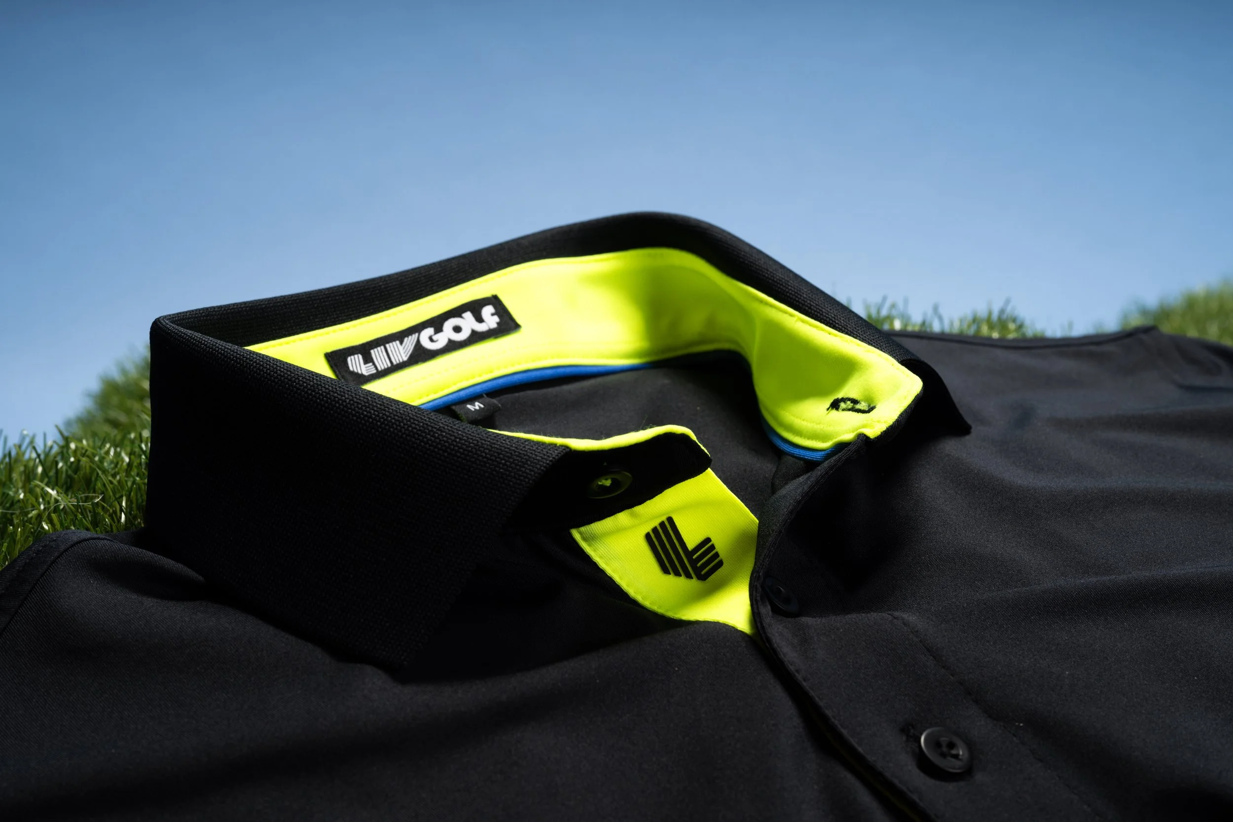 Close-up of a black golf bag with bright yellow and blue interior lining, lying on grass against a clear blue sky.