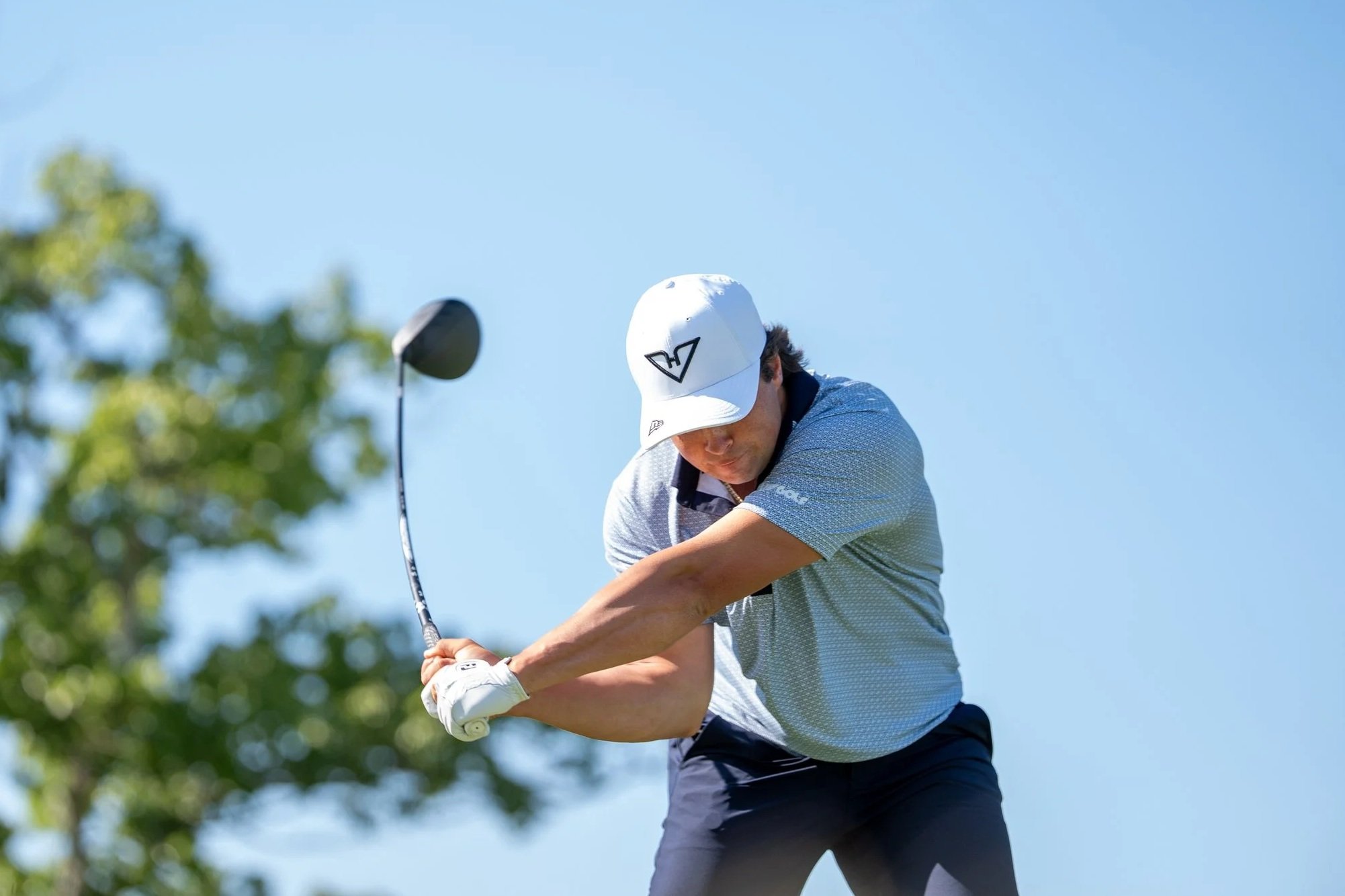 A man playing golf, swinging a golf club during daytime with a clear blue sky and trees in the background.