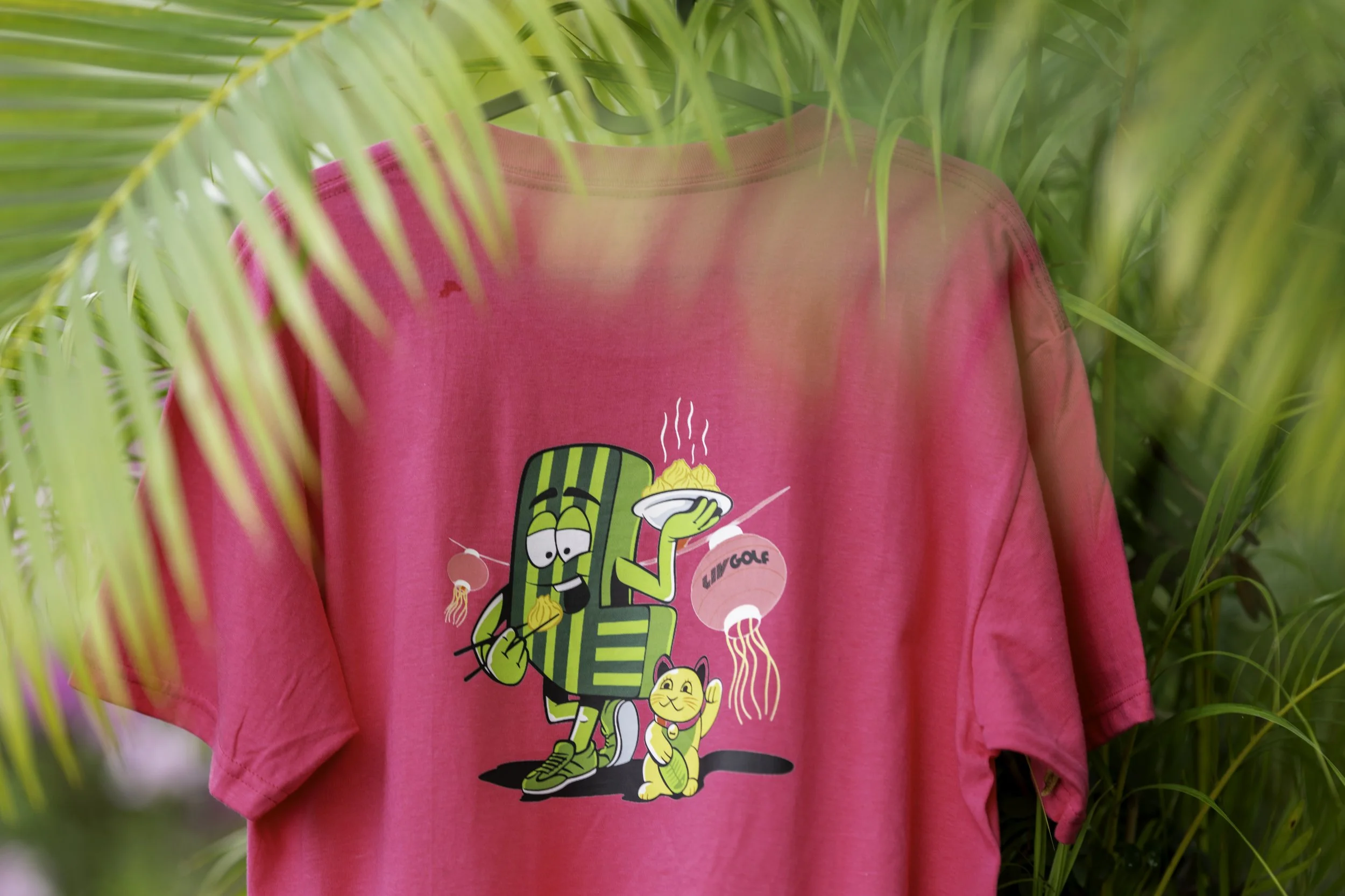 Close-up of a person wearing a bright pink sweatshirt with cartoon characters on it, surrounded by green tropical plants.