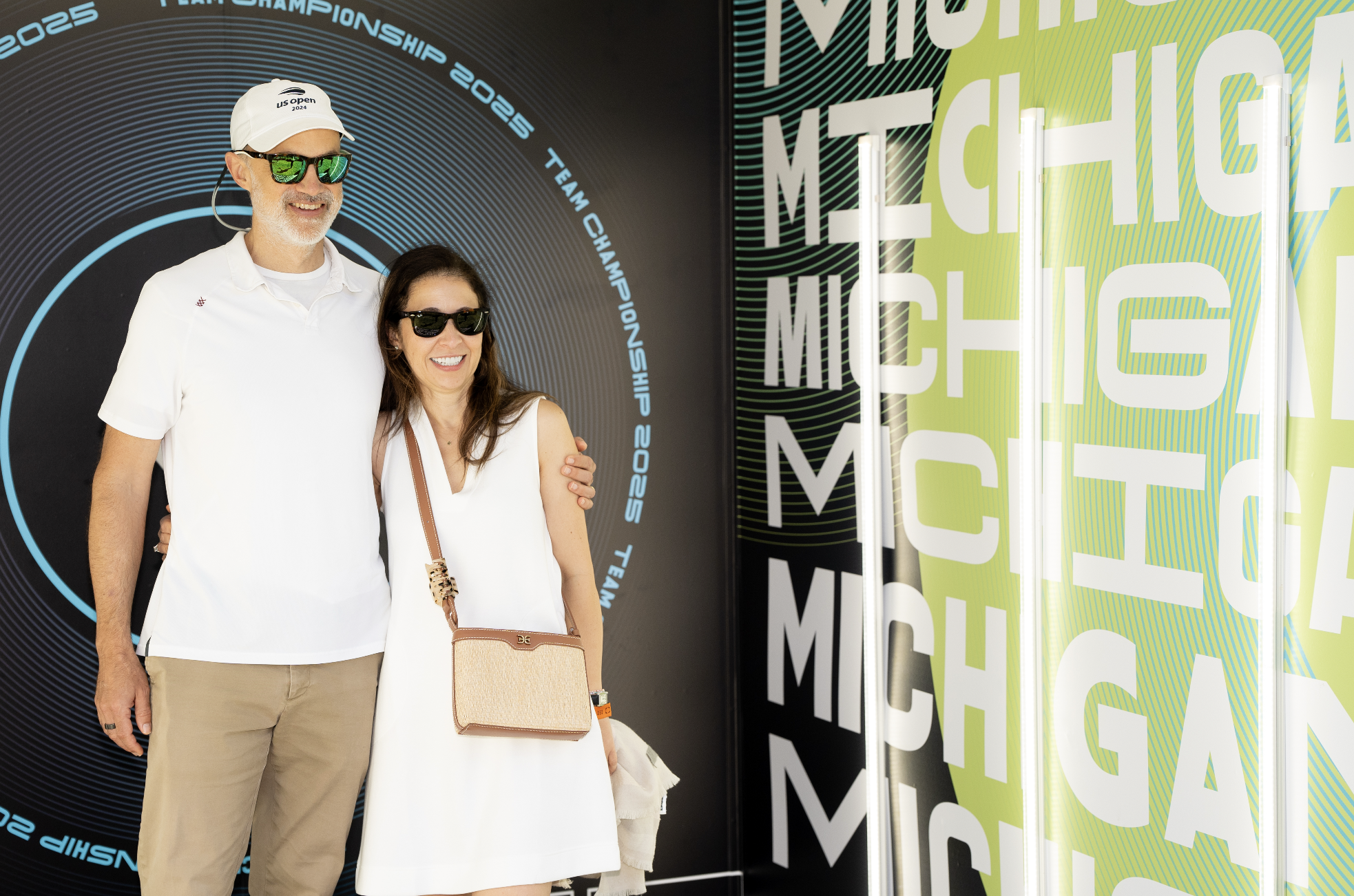 Two people, a man and a woman, posing together at a sports event. The man is wearing a white cap with the US Open 2024 logo, a white polo shirt, sunglasses, and beige pants. The woman is dressed in a white dress, sunglasses, and is carrying a beige handbag. They are standing in front of a colorful backdrop with large white letters and a circular design, indicating a tennis championship event.