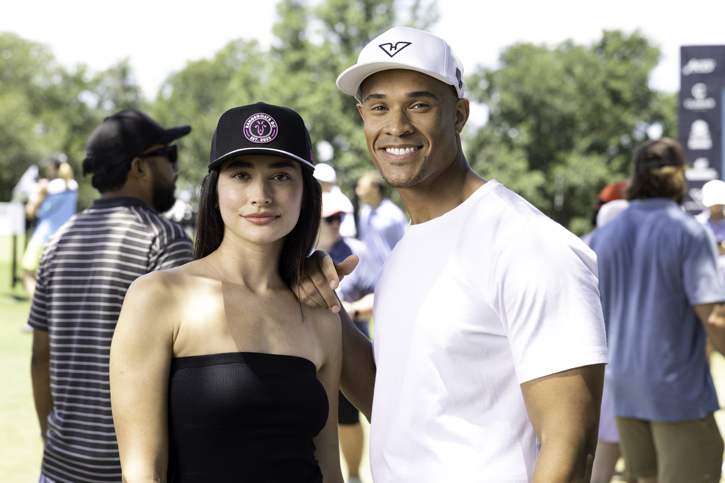 Two smiling people, a woman in a black strapless top and a man in a white t-shirt and cap, stand close together outdoors at a park or festival, with trees and other people in the background.