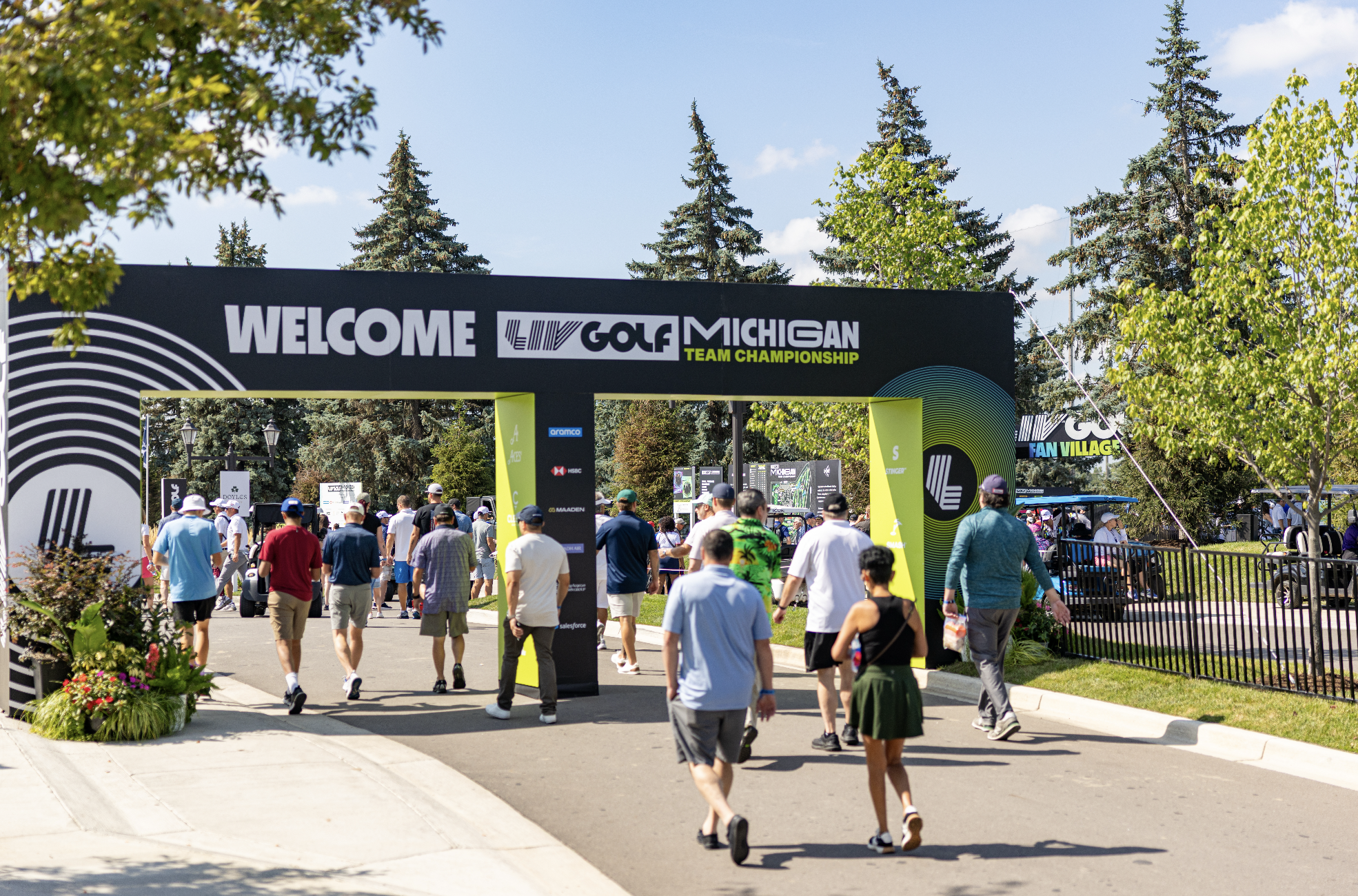 Crowds of people walking into the LIV Golf Michigan Team Championship event under a large black archway with signage. Trees and a partly cloudy blue sky are visible in the background.