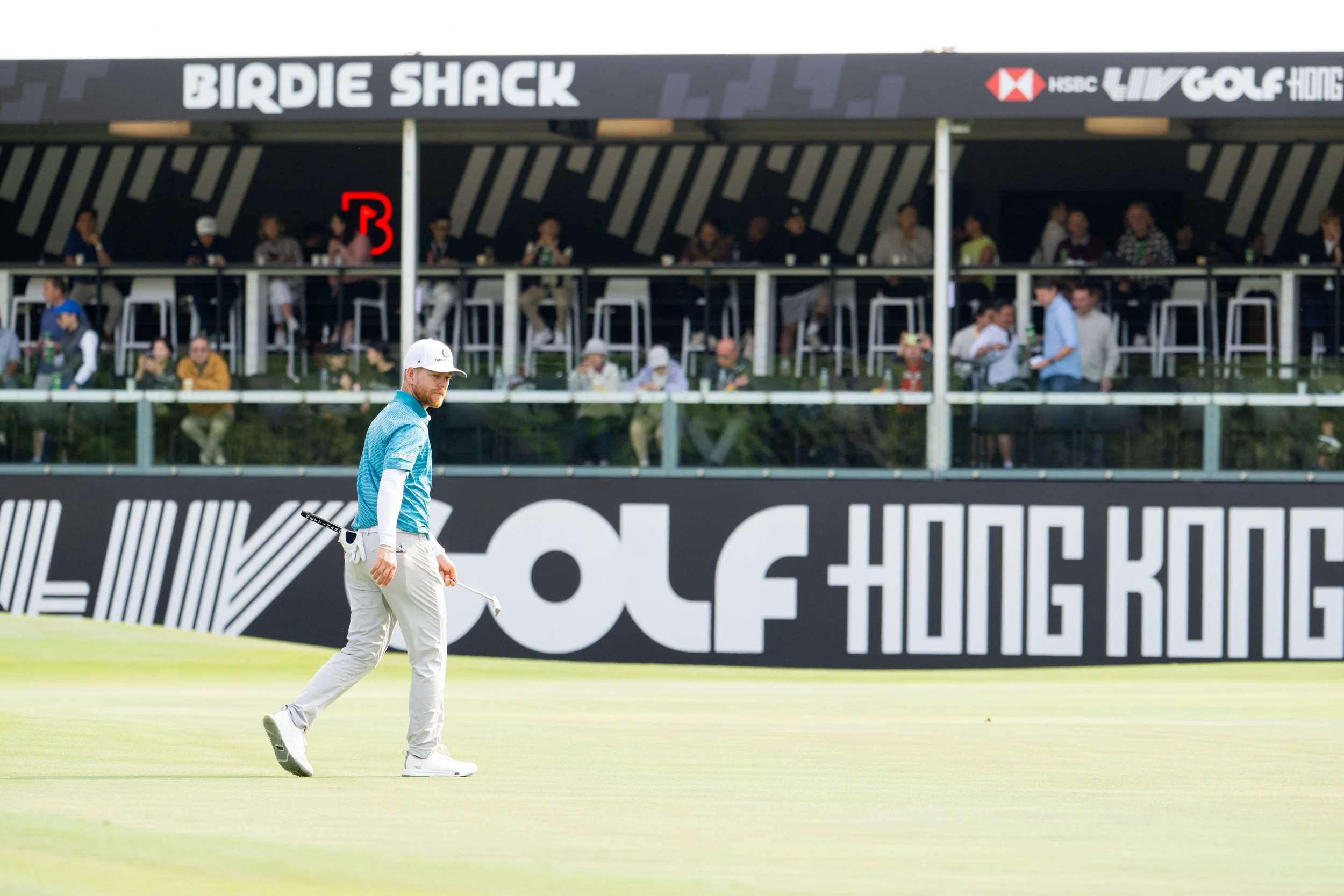 A golfer walking on a golf course with a putting green in the foreground and spectators watching from a shaded grandstand in the background during a professional golf tournament.