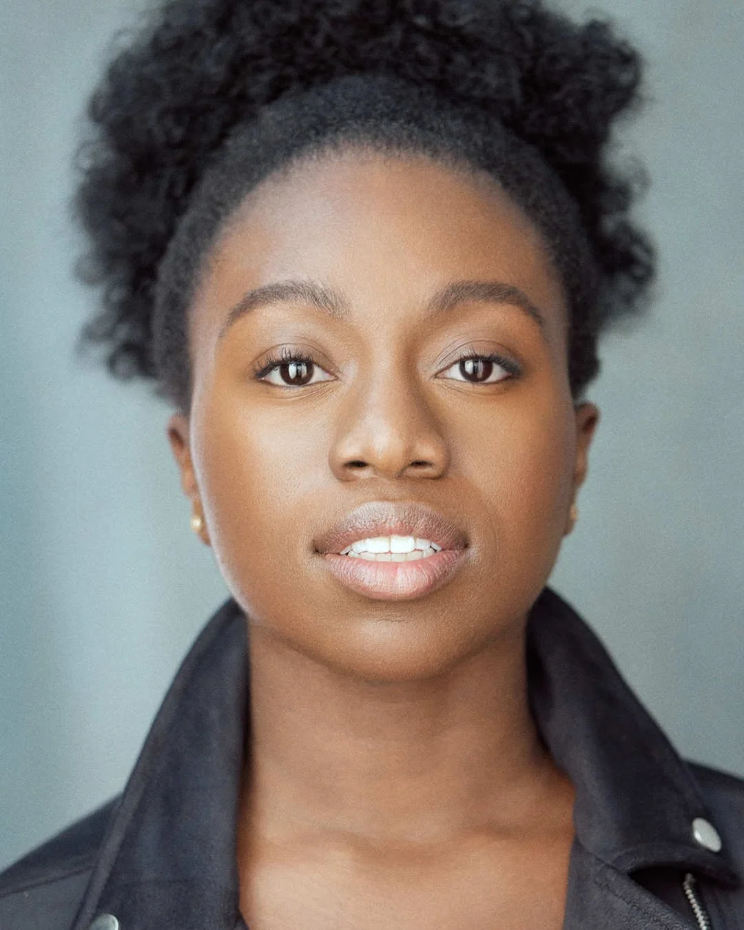 Close-up portrait of a young African American woman with natural hair and a black jacket, looking directly at the camera with a neutral expression.