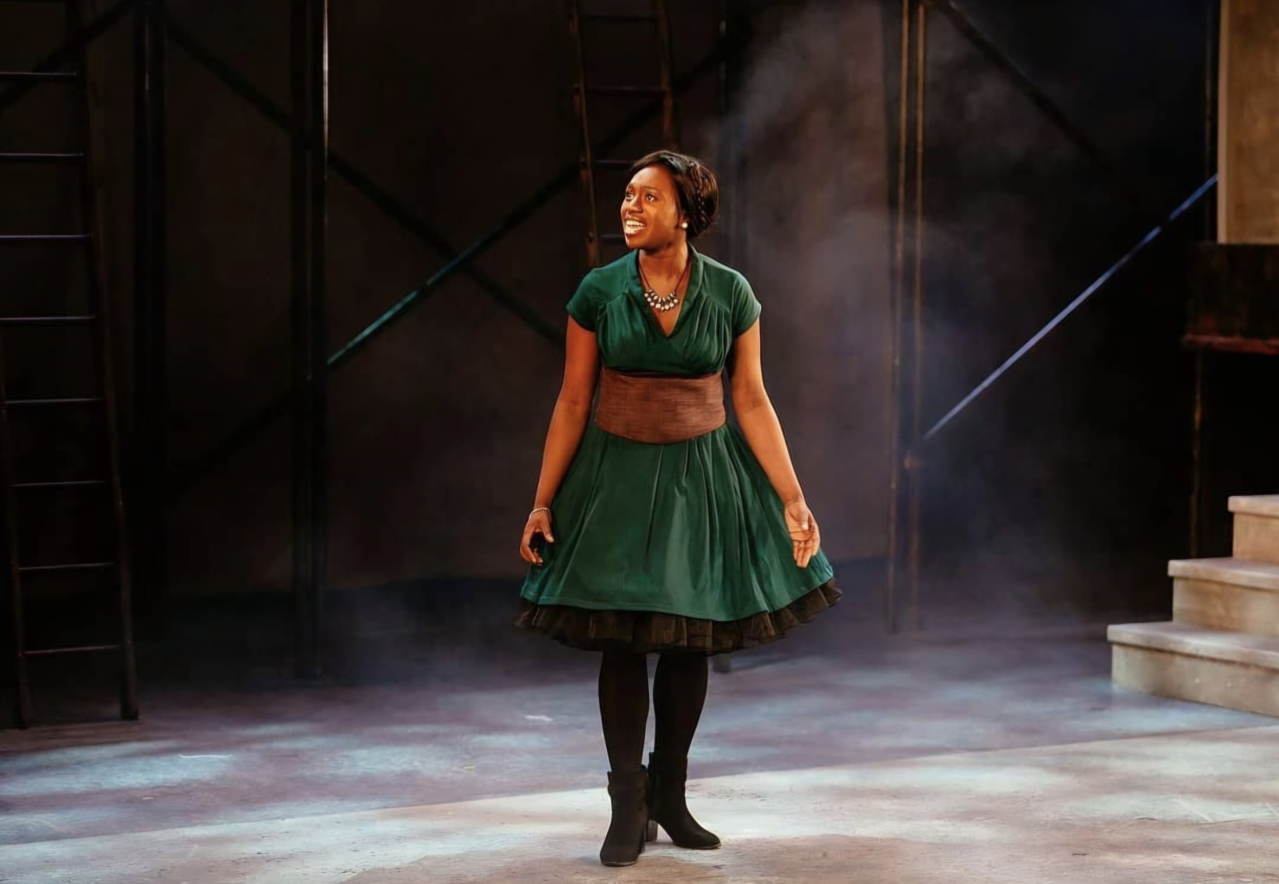 A woman performing on stage, wearing a green dress with a brown waist belt, black tights, black boots, and a necklace, standing in front of dark stage background with stairs on the right.